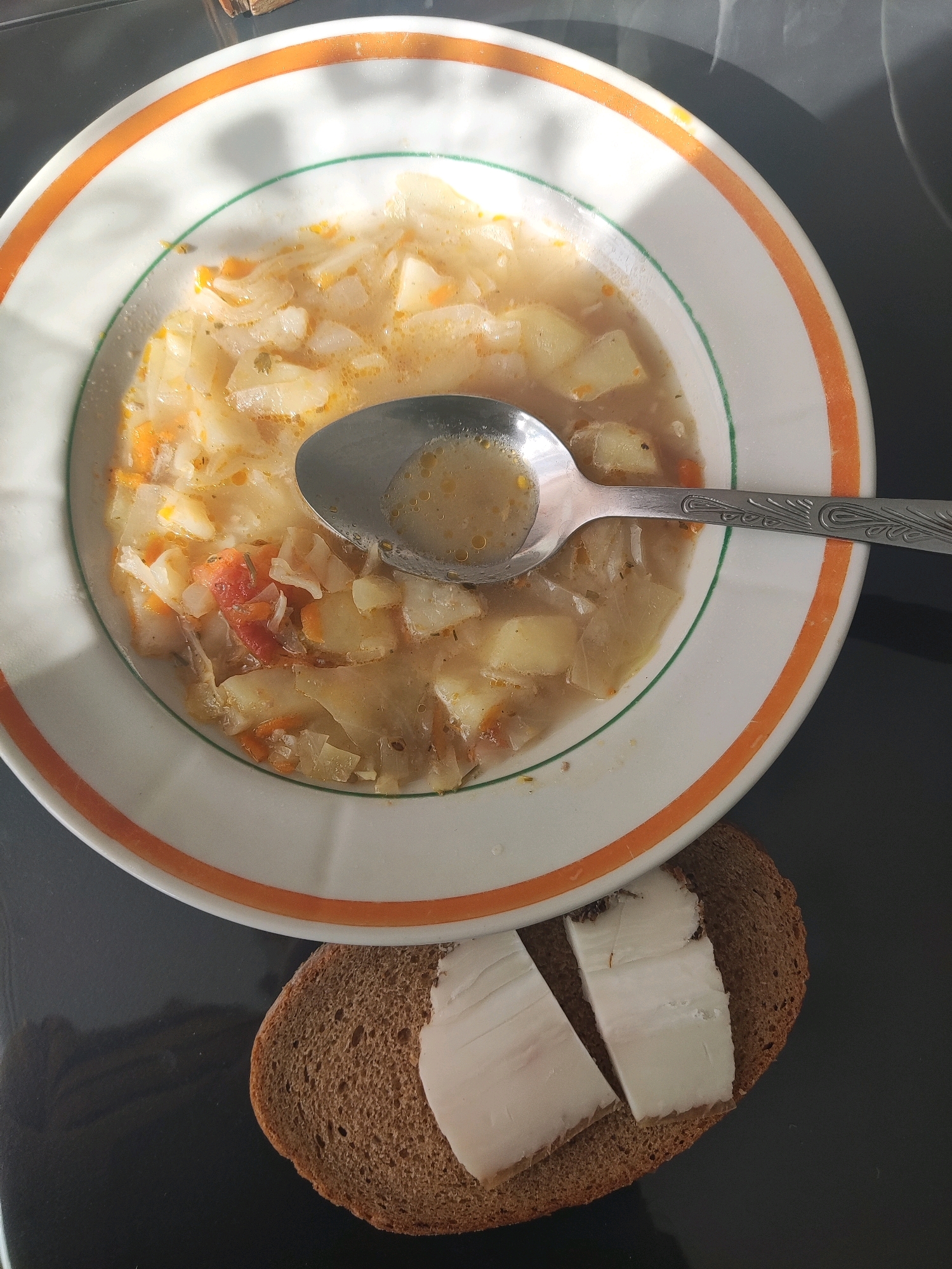 A close-up, overhead shot shows a bowl of vegetable soup and a slice of bread with lard. The soup is in a white bowl with an orange and green rim, and it contains pieces of potatoes, carrots, and possibly cabbage, with a silver spoon resting in it. The bread is dark rye, with two pieces of white lard on top. The scene appears to be set on a dark, reflective surface, possibly a table. The lighting suggests an indoor environment, with shadows cast on the soup bowl. No people or specific landmarks are visible, making it impossible to determine the exact location within Свіслацкі сельскі Савет, Belarus.