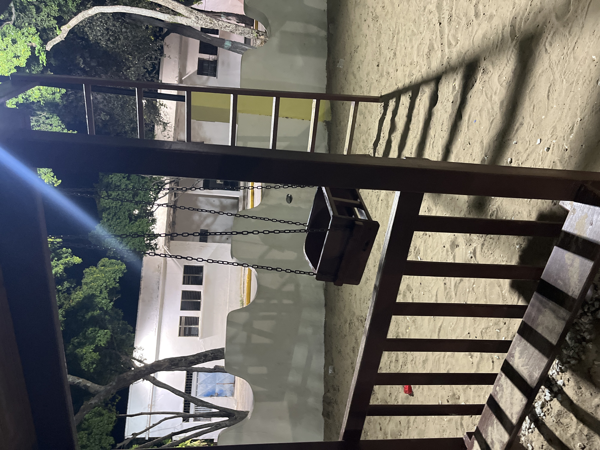 The image depicts a nighttime scene at a playground in Santa Ana, Venezuela. In the foreground, a wooden swing set is partially visible, with a dark wooden swing suspended by chains. To the right of the swing set, a ladder made of wood and metal rises vertically. The ground beneath the playground equipment is covered in sand, with some scattered pebbles and debris. In the background, a white building with multiple windows and a yellow-tiled section is visible. Trees with green foliage surround the building and playground. The scene is illuminated by artificial lighting, casting shadows across the sand and playground structures. There are no people visible in the image. A red toy is on the sand to the left.