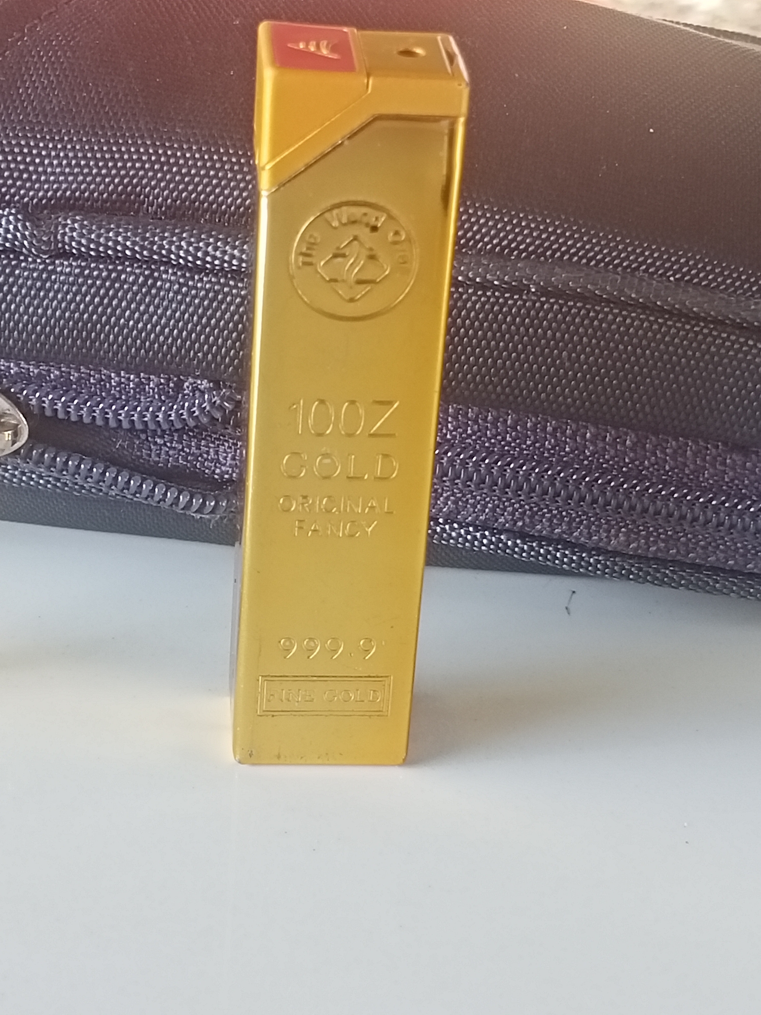 A gold-colored lighter, shaped like a gold bar, is the central object in the image. It is labeled "100Z GOLD ORIGINAL FANCY" and "999.9 FINE GOLD". The lighter has a small red section at the top and a circular emblem. The lighter stands on a white surface. Behind it is a dark-colored, textured bag or case with a zipper. The location or specific city in Indonesia is not evident from the image.