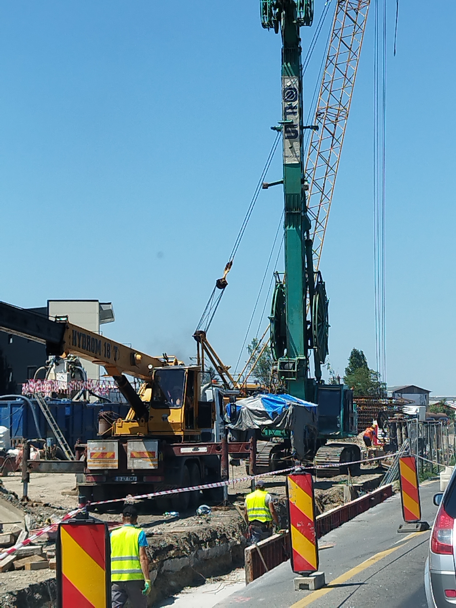 In Bragadiru, Romania, a construction site is active under a clear blue sky. Two large cranes dominate the scene. A yellow crane, identified by "HYDROM 18 T" on its boom, is positioned in the foreground. Behind it, a taller, green tracked crane with a latticed orange jib is engaged in work. Construction workers, some wearing high-visibility vests, are present on the site, with at least two visible. One worker in a yellow vest is walking away from the camera, while another in an orange vest is seen near the base of the green crane. The ground is disturbed with trenches and construction materials, including rebar. Safety barriers, consisting of red and white striped tape and yellow and red striped hazard posts, delineate areas of the site. A dark-colored car is partially visible in the lower right corner, suggesting the construction is adjacent to a road. The environment appears to be an urban or suburban area, with buildings visible in the background.