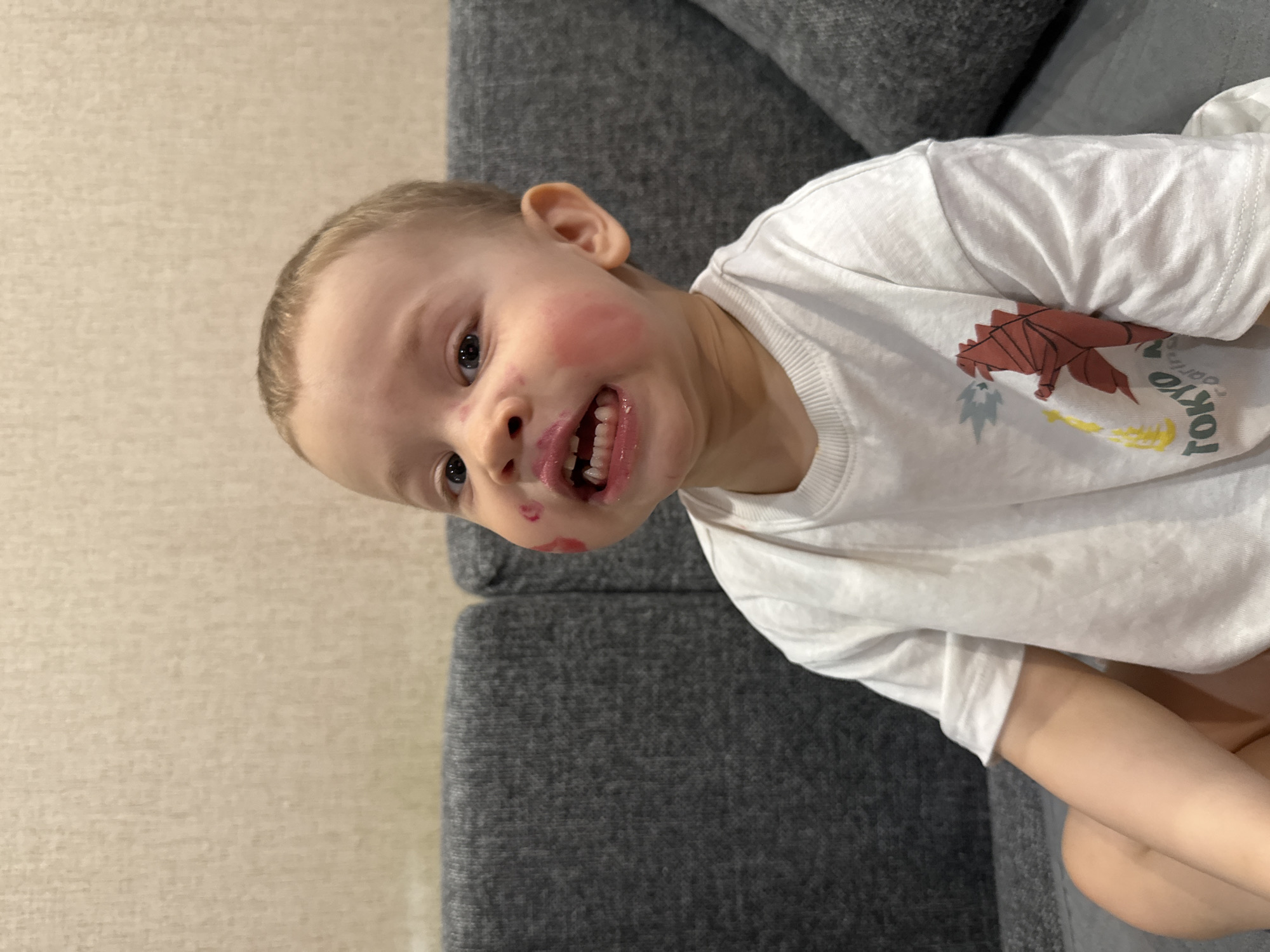 A young child with smeared red substance around the mouth and on the cheeks is seated on a gray sofa. The child is wearing a white t-shirt with a print on it. The background includes a textured beige wall. The location is not discernible as Rusanovo, Russia from the image alone.