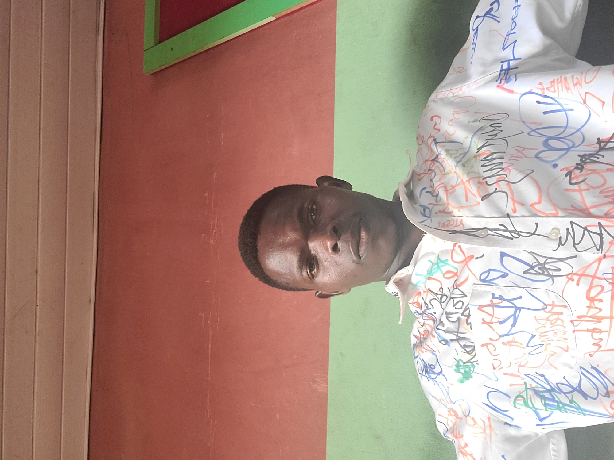 The image shows a close-up of a young Black male wearing a white button-down shirt covered in colorful graffiti. The subject is looking directly at the camera with a neutral expression. The background is divided into two distinct sections: on the left, a reddish-brown wall, and on the right, a light green wall. The scene appears to be outdoors, possibly in a courtyard or public space, given the contrast in wall colors. The lighting suggests it could be daytime, with no clear indication of the specific time. The man's shirt is a focal point due to the vibrant and chaotic nature of the graffiti, which obscures the fabric pattern and details of the garment itself. Some of the graffiti appears to be in English or a similar Latin-based script, while other markings are abstract doodles. The overall impression is one of a casual setting, possibly in Nigeria, as indicated by the location context. The image is taken from a slightly tilted angle.