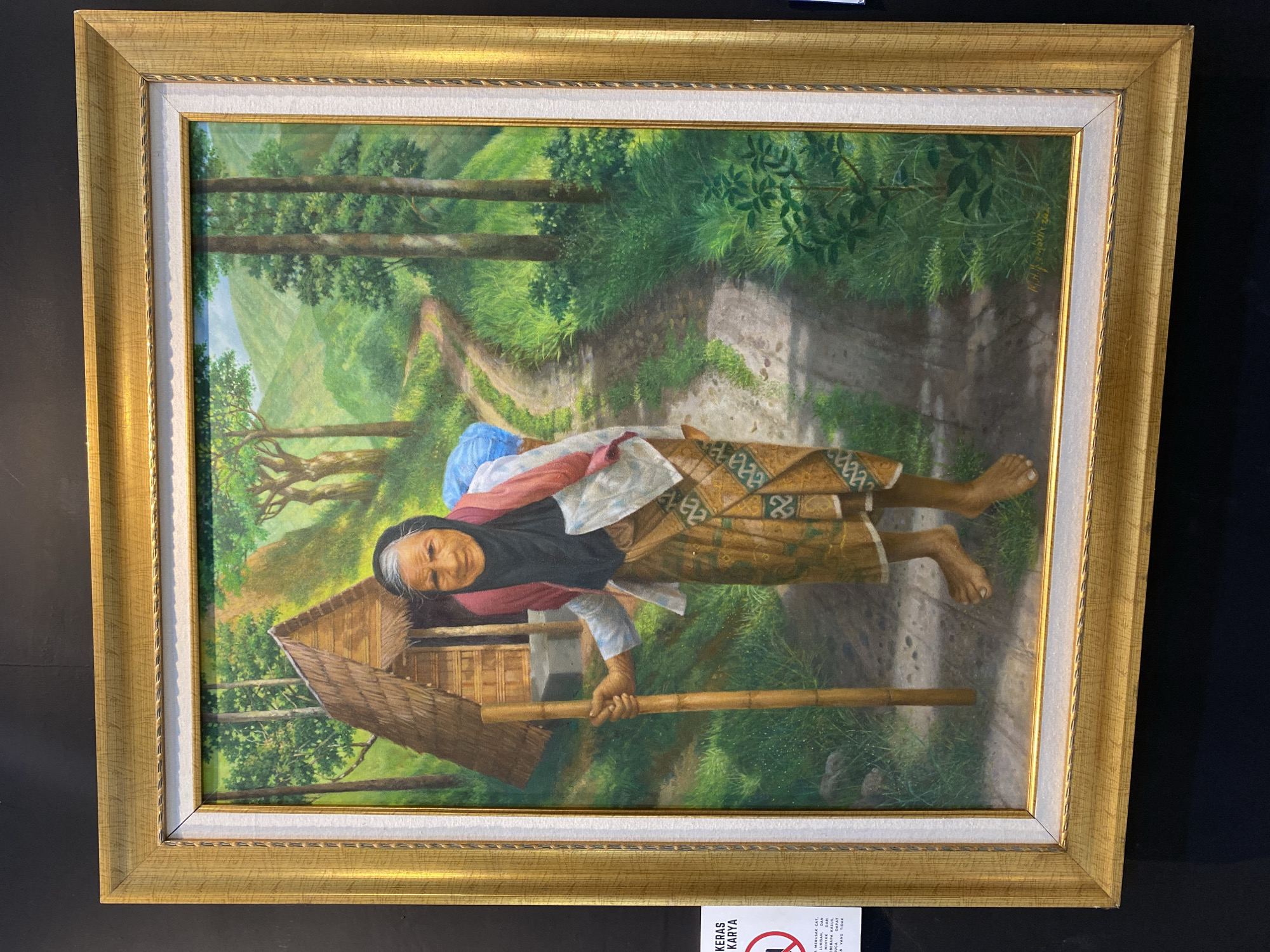 The image is a medium shot of a framed oil painting displayed against a black background. The painting depicts an elderly Indonesian woman with grey hair, wearing a black headscarf and a pink shawl over a white shirt and a patterned sarong. She is barefoot and holding a wooden staff, walking along a dirt path in a lush, green, hilly landscape. To her left is a small, rustic hut with a thatched roof. The background shows a winding path, trees, and rolling hills under a blue sky. The painting is in a golden, ornate frame. A white informational sign with red text and a red circular "no" symbol is partially visible on the left side of the frame. The signature "K.H. Sastrawijaya 2007" is present on the bottom right of the painting.