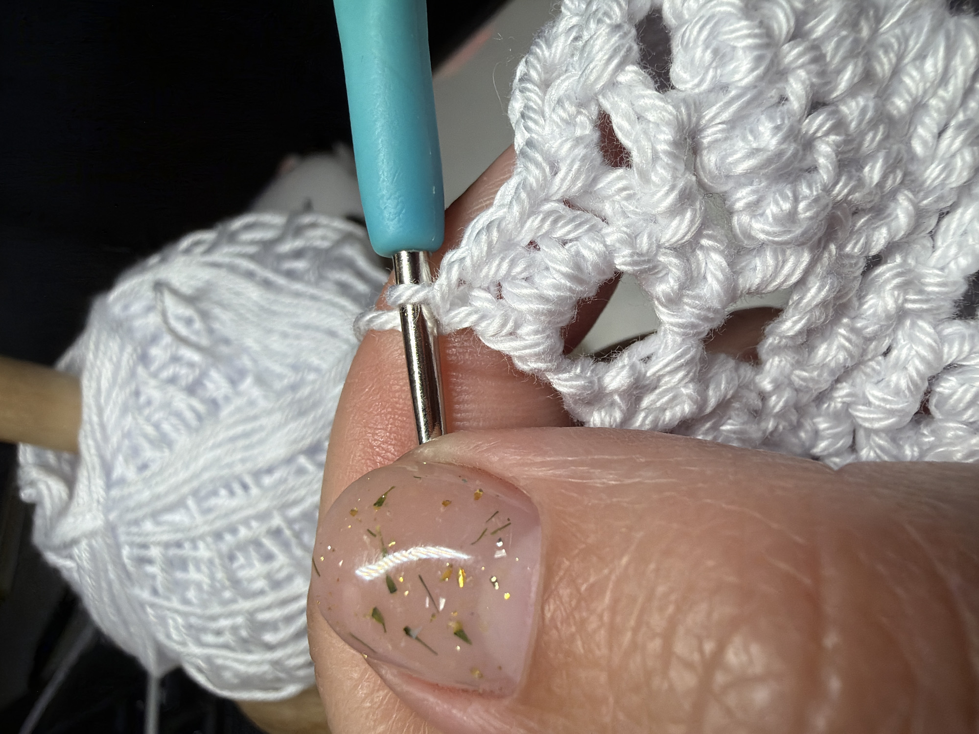 A close-up shot shows a person crocheting with white yarn using a light blue and silver crochet hook. The finished crochet work is white and lacy. In the background, there is a ball of white yarn. The person's thumb is visible, with a light pink nail covered in flecks of gold and green. There is no indication that the image was taken in Chaikovsky, Russia.