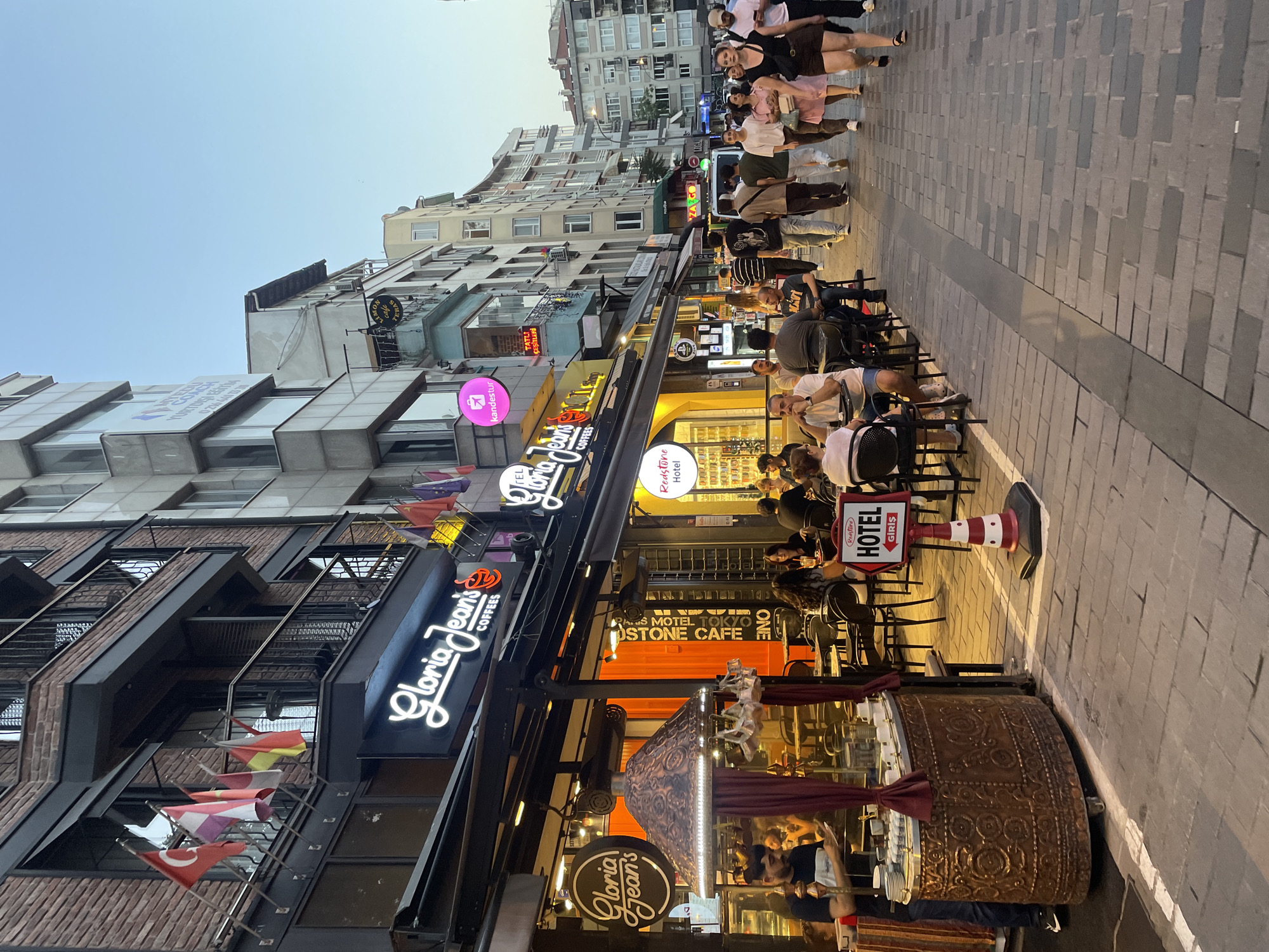The image depicts a bustling pedestrian street in Kadıköy, Turkey, likely during the late afternoon or early evening, with the sky showing a clear, soft blue hue. The street is lined with multi-story buildings featuring red brick facades on the left and lighter colored, modern structures on the right. Numerous flags are displayed on the buildings, including the Turkish flag and several European country flags.
On the left side of the street, a cafe with outdoor seating is prominent. The cafe's awning extends over a seating area where several people are seated at tables. The cafe's signage clearly reads "Gloria Jean's COFFEES." A large, ornate, copper-colored display with a decorative lamp hangs over a counter within the cafe. A man in a blue shirt and jeans is standing behind the counter, appearing to serve customers.
Further down the street, more cafes and shops are visible, with more outdoor seating areas occupied by patrons. People are walking along the sidewalk, some in groups and others individually. A red and white striped signpost with a "HOTEL" symbol and an arrow pointing to the right is visible in the middle ground. The street is paved with grey and darker grey interlocking rectangular bricks. The overall atmosphere suggests a vibrant urban commercial area.