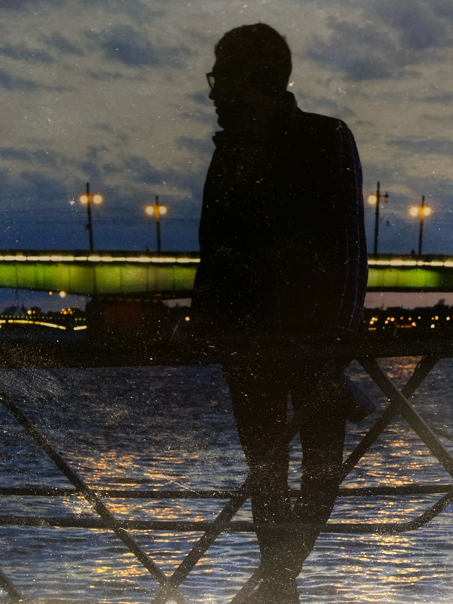 The image shows a silhouette of a person standing on a railing overlooking a body of water. The person is wearing glasses and a jacket. The background features a bridge with green lights and streetlights, indicating an urban setting. The time of day appears to be dusk or evening, with a cloudy sky and reflections of lights on the water. The location is identified as Zarechny, Russia. The water is rippling, and the overall image has a somewhat grainy or textured appearance, possibly due to the photograph's age or the way it was captured.