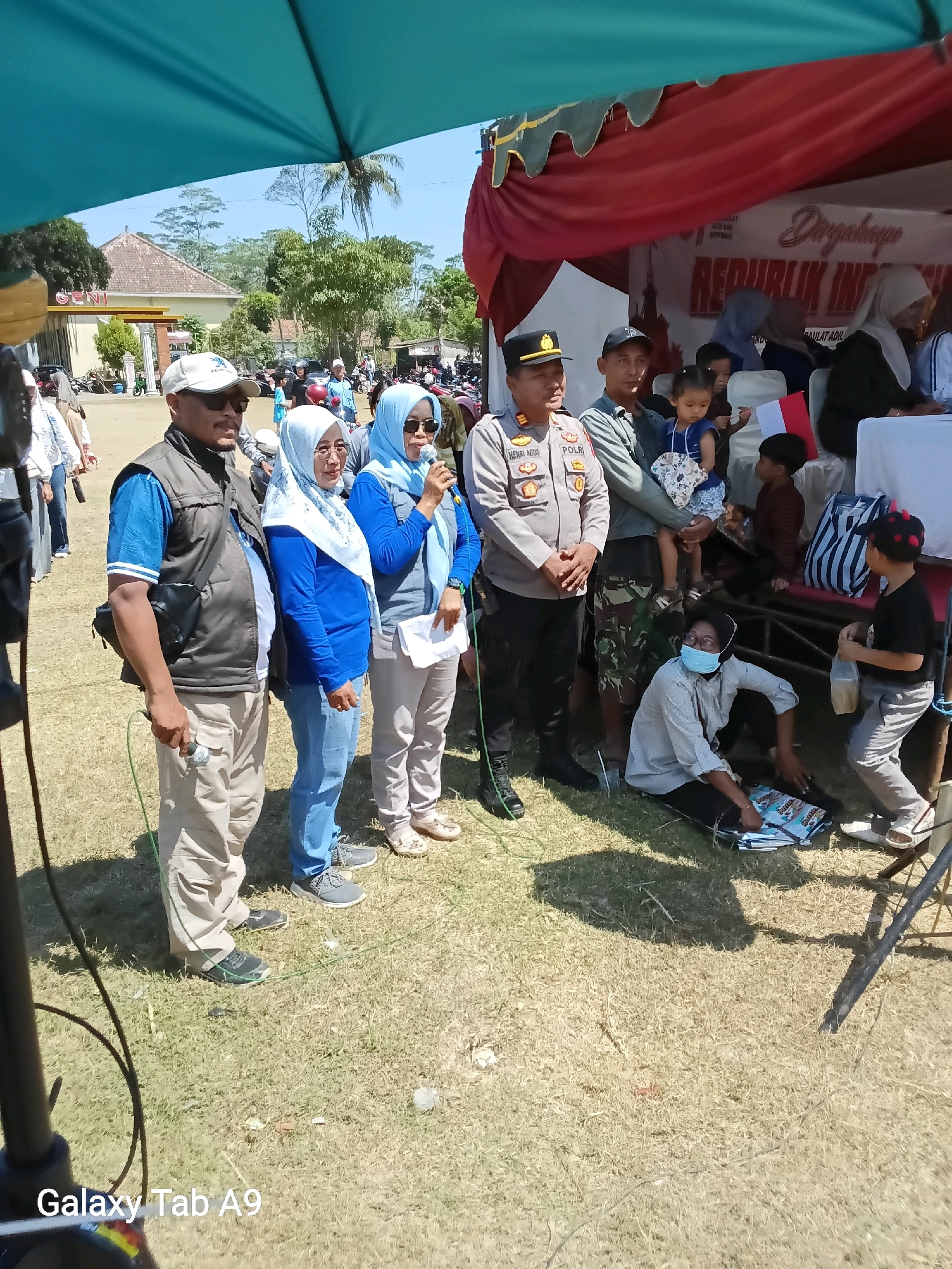 In Ngadirenggo, Indonesia, an outdoor event is taking place under a blue tarp. A group of people are gathered, including several women in hijabs and a man in a police uniform. The police officer, identified as HENRI AGUS, is wearing a grey uniform with "POLRI" visible on his jacket. He is standing to the left of the frame, with his hands clasped. Next to him, a man in a dark vest and a blue shirt is holding a microphone. Further to the right, a woman in a blue hijab is also speaking into a microphone. Behind them, a red banner with white text that includes "Dirgahayu REPUBLIK INDONESIA" is visible. Several children are present; one girl in a blue dress is being held by a man, and another child in a striped shirt is seated. A person wearing a light-colored shirt and a face mask is seated on the ground in the lower right quadrant of the image, with papers spread out in front of them. The ground is covered in dry grass, and in the background, buildings and trees are visible, suggesting a rural or semi-rural setting. The overall scene appears to be a public gathering or ceremony.