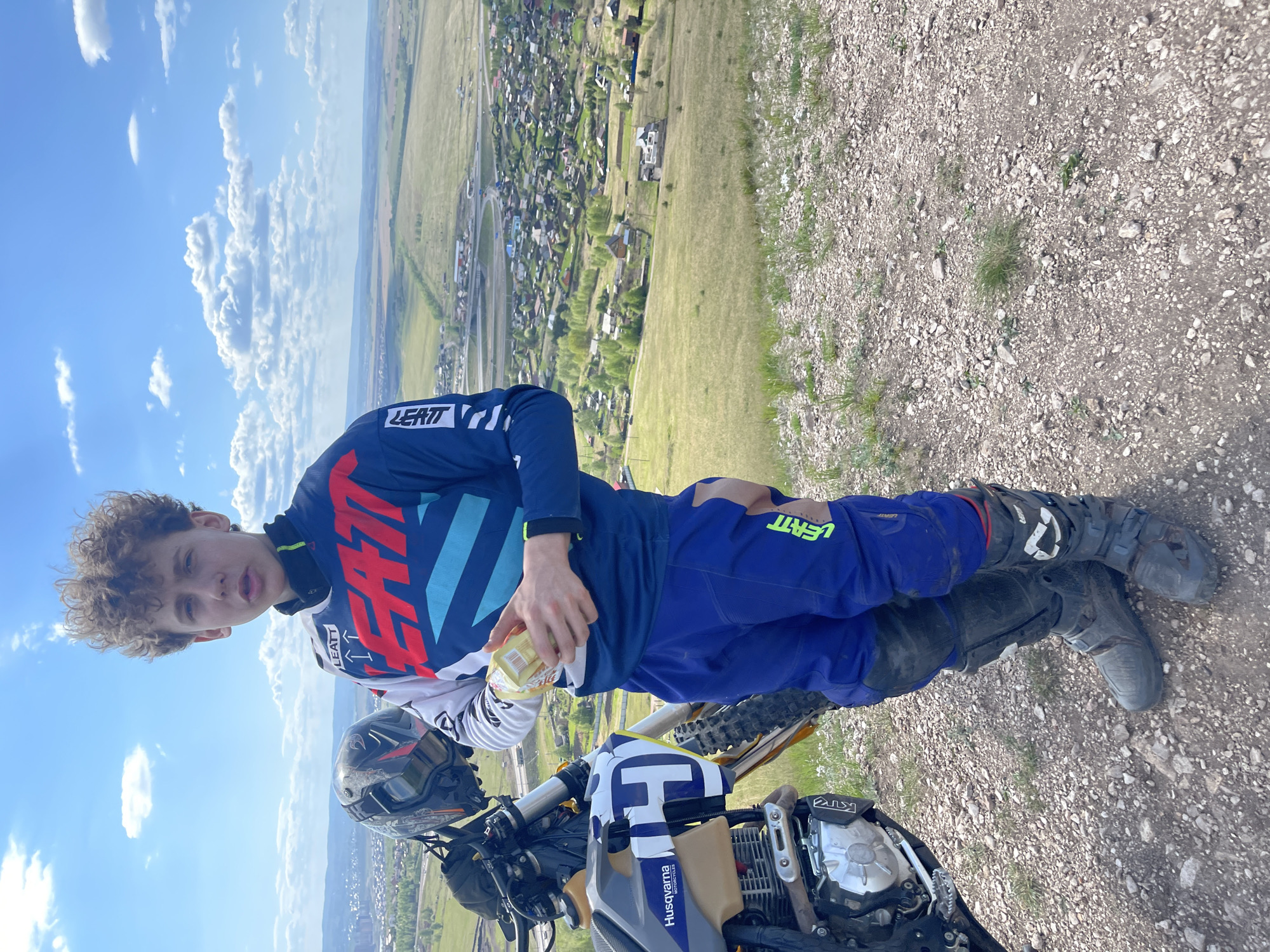 A young man, presumably a teenager, is standing with a dirt bike on a rocky, grassy hillside overlooking the cityscape of Солонцы, Russia. He is wearing a blue, white, and red long-sleeved racing jersey with "LEATT" printed on it, along with matching blue pants and grey motocross boots with knee pads. He holds a small clear bottle, likely containing a beverage. To his right stands a dirt bike, facing towards the viewer, featuring a grey and blue color scheme with the word "Husqvarna" visible on the side panel and a prominent white "H" on the front. The dirt bike's helmet is visible on the handlebars. The background features a sprawling landscape with a clear sky filled with puffy clouds above the town of Солонцы. The ground is made of loose gravel.