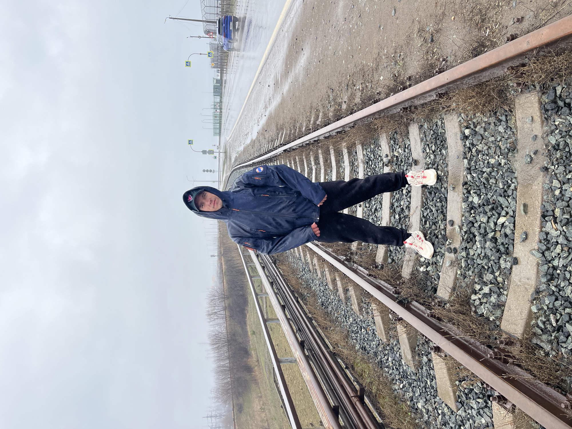 An outdoor, full-body shot depicts a person standing on railway tracks on an overcast day in Chaikovsky, Russia. The individual, appearing male, is positioned centrally, facing the camera with a neutral expression. They are wearing a dark blue hooded jacket, dark pants, and light-colored athletic shoes with red accents. A dark cap is visible under the hood. A patch is on the left arm of the jacket.
The subject stands with legs spread wide, one foot on a concrete sleeper and the other on the gravel ballast next to the rail. The railway tracks, made of metal rails on concrete sleepers and grey gravel ballast, extend into the distance. Sparse, dry vegetation grows between the sleepers and rails.
To the right of the tracks, a wet, dirt or paved area runs parallel, leading to a road in the distance where a blue car is visible, creating a spray of water, indicating recent rain or wet conditions. Several yellow diamond-shaped road signs are also seen along this area. To the left of the tracks, another set of rails runs parallel, beyond which are bare trees and utility poles under a muted, light grey sky. The overall weather appears cold and damp, with the ground visibly wet. The time of day is daytime, but with heavy cloud cover. No legible text is visible in the image.