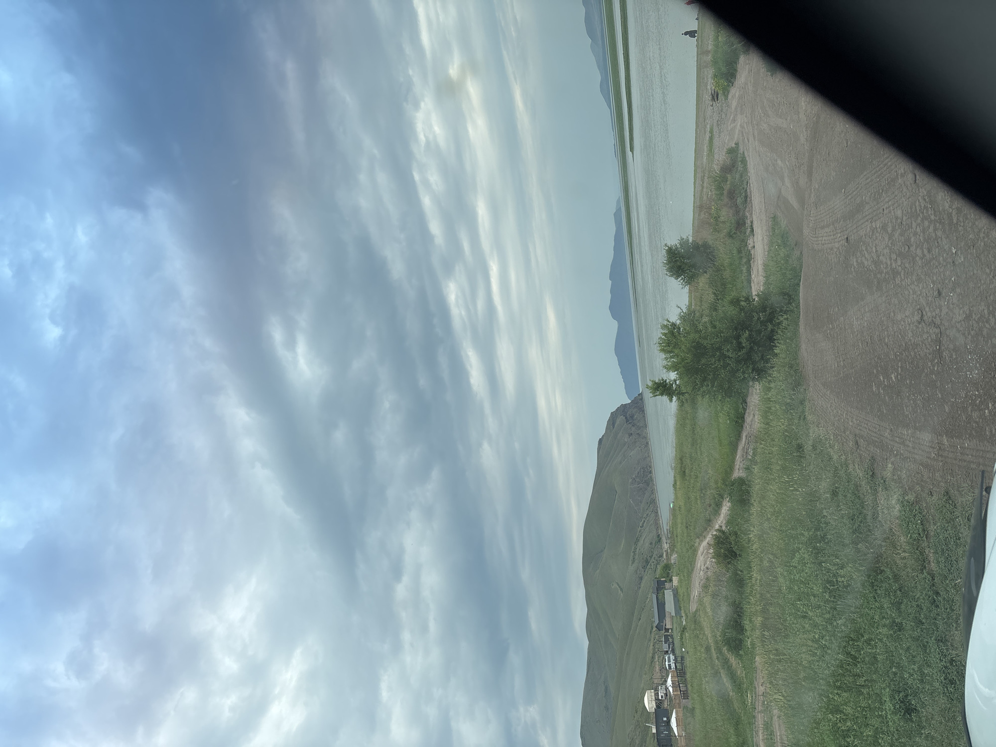 The image depicts a coastal landscape with rolling green hills meeting a wide expanse of water under a cloudy sky. In the foreground, a dirt road leads towards the water, with tire tracks visible on the dusty surface. To the left of the road, a small settlement with several structures, including white domed tents and dark-colored buildings, is visible amidst lush green grass. A few vehicles, including a white van, are parked near the settlement. The water stretches out to the horizon, where more distant hills or landmasses are faintly visible. The sky is filled with dramatic, layered clouds, suggesting overcast or pre-storm conditions. The scene appears to be a remote, natural setting, possibly a campground or a small village near a body of water, with no specific identifiable landmarks pointing to Ust-Abakan, Russia.
