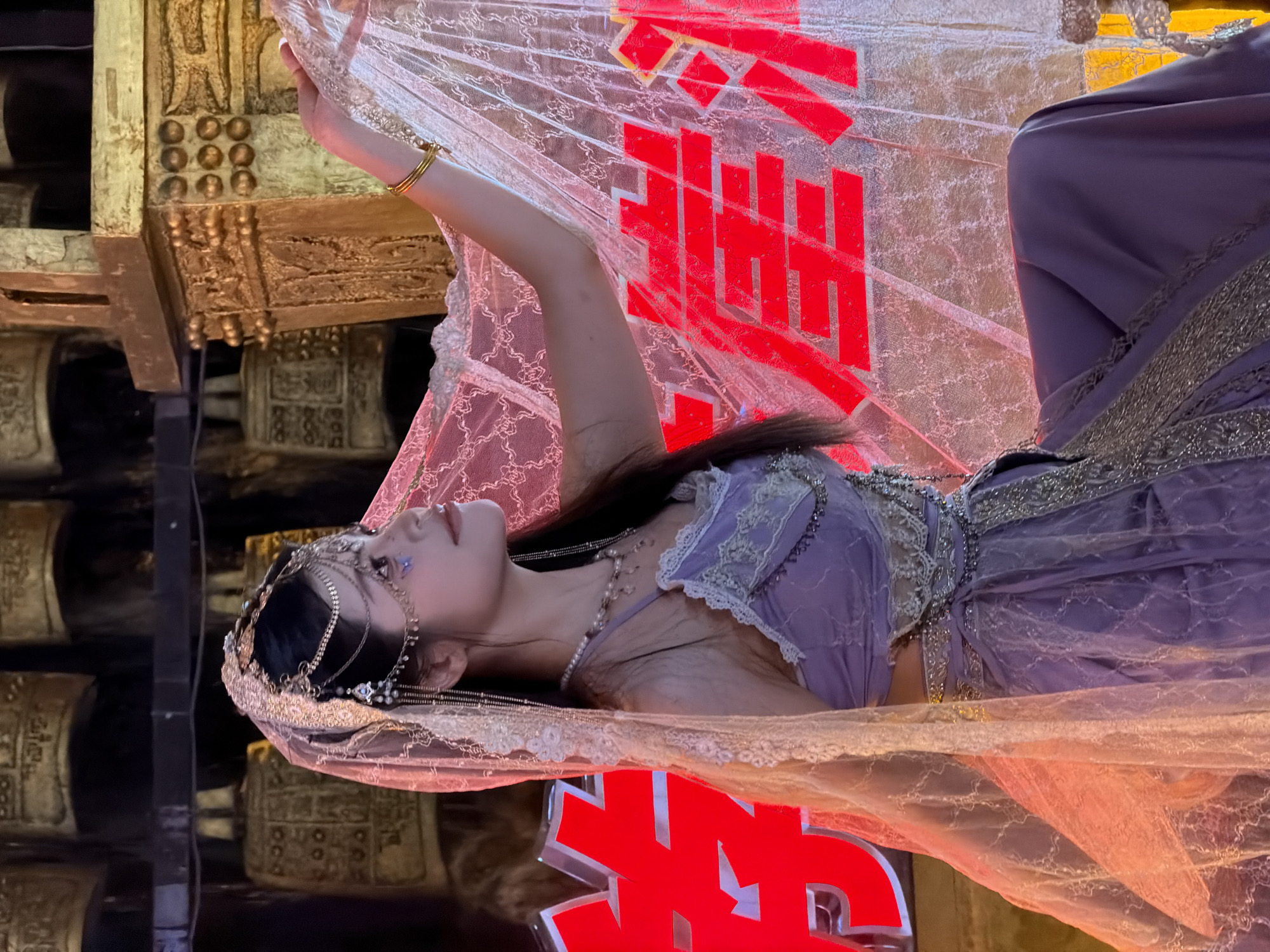 This image shows a woman in a traditional Chinese-inspired outfit, possibly performing or posing for a photo. She is wearing a flowing purple garment with intricate gold detailing and a sheer, lacy veil. Her hair is adorned with a jeweled headdress. She is positioned to the left of the frame, with her arm raised, and her face is turned upwards. The background features elements that suggest a theatrical or ceremonial setting. To the left, there are large, ornate gold objects that resemble bells or gongs, stacked vertically. To the right and behind the woman, a large, bright red Chinese character is visible, illuminated by light. The overall scene has a dramatic and artistic feel, with the red character and gold elements creating a strong visual contrast with the woman's purple attire. The lighting appears to be artificial, suggesting an indoor or nighttime event. The presence of the Chinese character and the attire suggest a connection to Chinese culture or tradition.