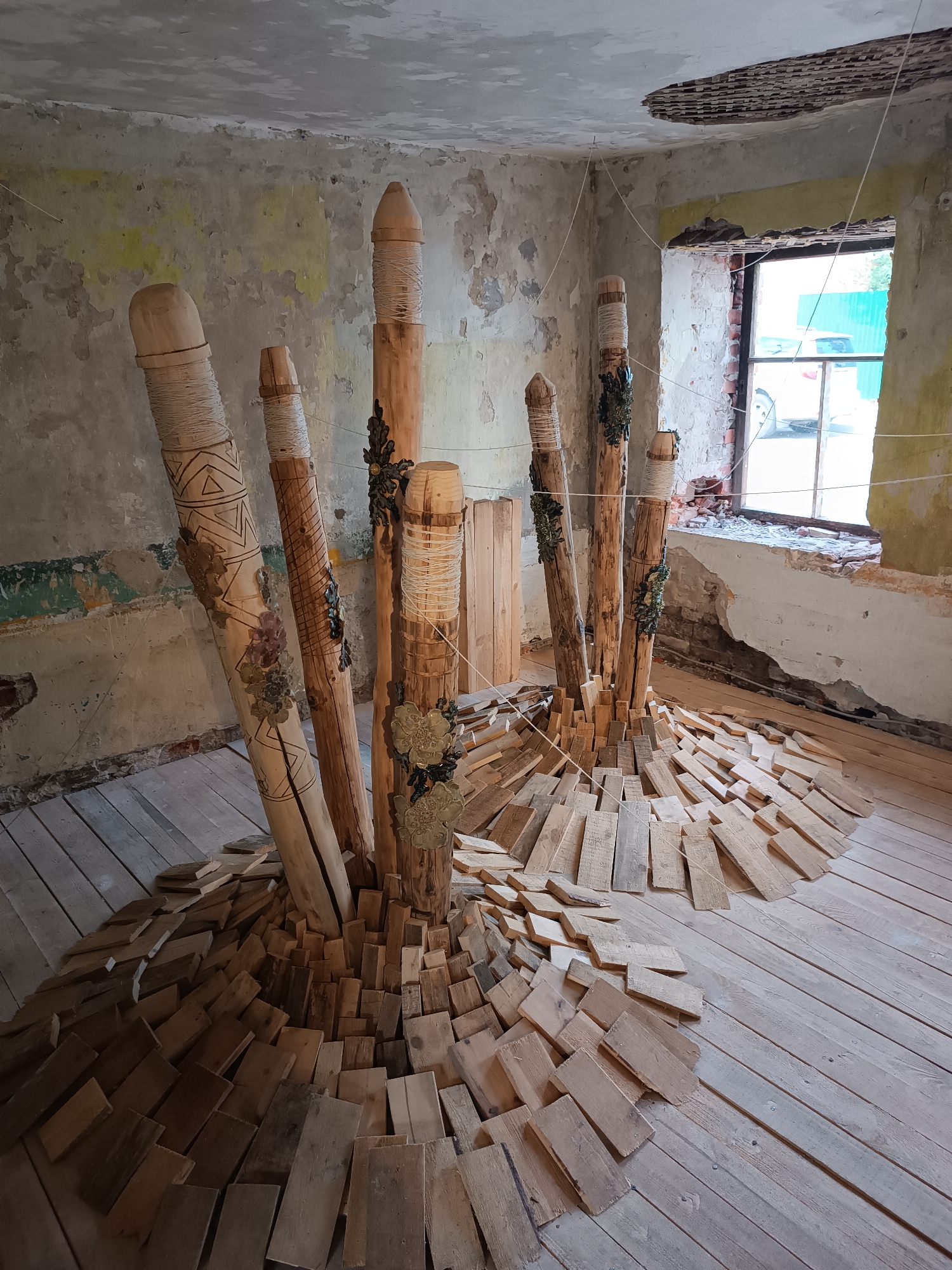 This image captures an interior space in Yelets, Russia, featuring a sculptural installation. The installation consists of several tall, wooden posts of varying heights and thicknesses, some of which are carved with geometric patterns and wrapped with white twine. Decorative elements, resembling clusters of stylized flowers or leaves, are attached to some of the posts. The base of the sculpture is surrounded by a circular arrangement of numerous small, light-colored wooden planks laid on the floor. The environment appears to be an unfinished or derelict room, with rough, textured walls showing peeling paint and plaster. A large, somewhat broken window on the right side of the frame reveals an outdoor scene with a fence and a vehicle. The floor is made of aged wooden planks. The overall impression is one of an art installation within a decaying architectural setting.