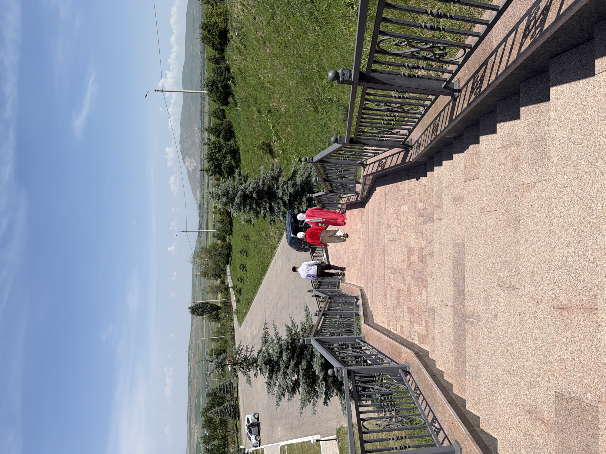 In this outdoor vertical shot, three people ascend a wide stone staircase with decorative metal railings, on a bright, sunny day. The staircase is lined with pinkish-tan stone pavers and dark metal railings with intricate scrollwork. The group consists of two women in bright red traditional clothing, one with a white headscarf, and a man in a white shirt and dark pants. They are walking away from the camera towards a paved area where a black SUV is parked.
Beyond the staircase, a paved road curves through a landscape of rolling green hills and scattered trees. In the distance, a range of mountains is visible under a clear blue sky with scattered white clouds. A white car is parked on the left side of the paved road, near a manicured area with evergreen trees. The overall scene suggests a tranquil, scenic location possibly a park or a viewpoint. The shadows on the staircase indicate the sun is high in the sky.