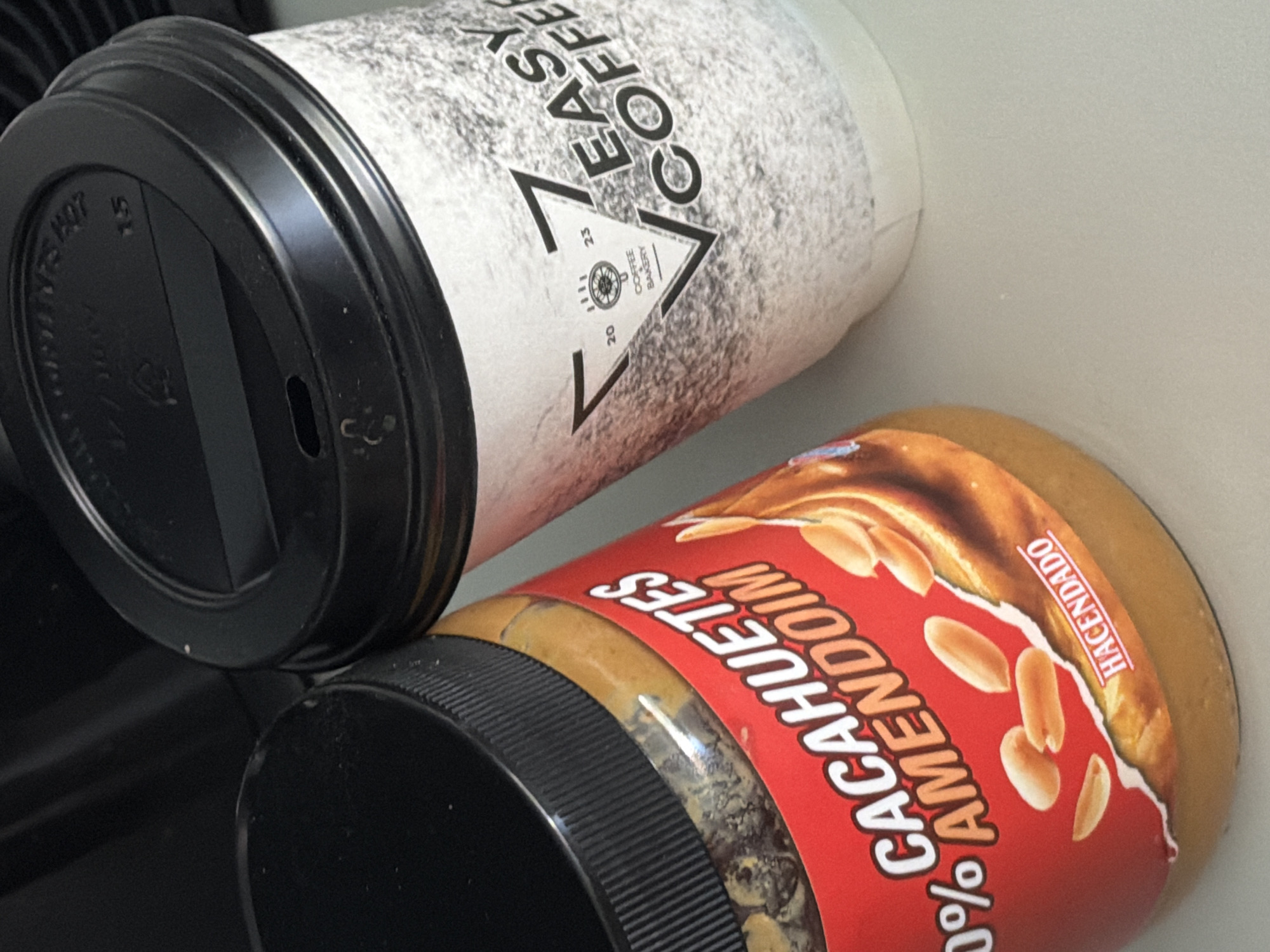 The image shows a close-up view of a disposable coffee cup and a jar of peanut butter, likely on a flat surface. The coffee cup has a black lid and a white paper sleeve with black text. The text on the sleeve reads "EASY COFFEE" and features a stylized logo with the words "COFFEE BAKERY". There are also numbers "20", "23", and a wheel graphic. The coffee cup is tilted and appears to be made of paper, with a rough, textured surface.
The jar of peanut butter has a red label with white text that reads "CAHUE TES" and "AMEND OIM". There is also text in a smaller font that says "HACENDADO". Images of peanuts are visible on the label, and the jar has a black lid with a ribbed texture. The peanut butter itself is a light brown color and is visible through the jar, with some texture from the peanuts. The overall lighting suggests it is daytime, but no specific time can be determined. The scene is simple and domestic, with no discernible weather or strong emotional cues. There are no people visible in the image. The specific location of Finestrat, Spain is provided context but is not directly visible in the image itself.