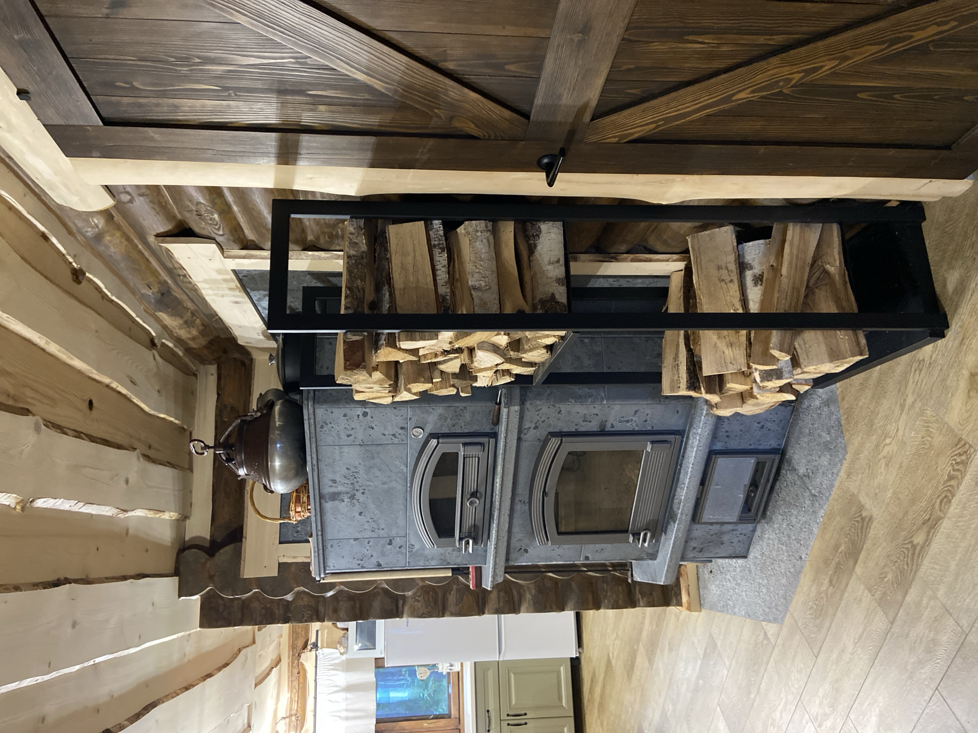 The image shows an interior scene of a rustic room, possibly a kitchen or living area, with prominent log walls and a wooden beam ceiling. In the foreground, a large, dark grey tiled stove is the central feature, with two visible firebox doors, one above the other. To the right of the stove is a black metal rack filled with neatly stacked firewood. Adjacent to the firewood rack and the stove is a dark brown wooden barn-style door with diagonal planks and a black handle. In the background, a white refrigerator and kitchen cabinets with green doors are visible through an opening, along with a window looking out onto a green landscape. The flooring appears to be light-colored wood or laminate. The overall aesthetic suggests a cozy, traditional, or country-style dwelling. The location of Забегаево, Russia is not visually represented in the image.