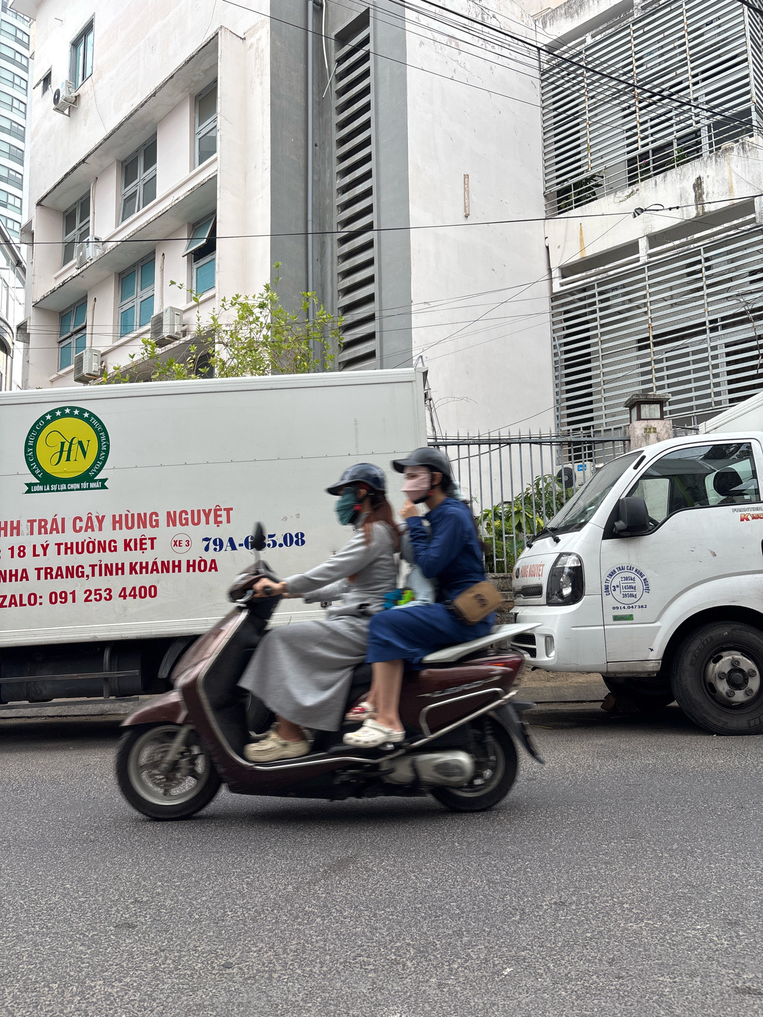 This is a medium shot, eye-level, outdoor, daytime, urban scene in Nha Trang, Vietnam. The primary focus is a woman riding a scooter, with another person behind her, both wearing face masks and modest clothing. They are moving from left to right in front of a white truck. The truck has Vietnamese text on its side, indicating it belongs to a fruit company and lists an address on Lý Thường Kiệt street in Nha Trang, Khanh Hoa province, along with a phone number. To the right of the truck, part of a white van is visible. In the background, there are buildings with multiple windows and horizontal blinds, and overhead power lines are prominent. The road surface is asphalt. The overall impression is of a typical street scene in a Vietnamese city, with traffic and commercial vehicles.