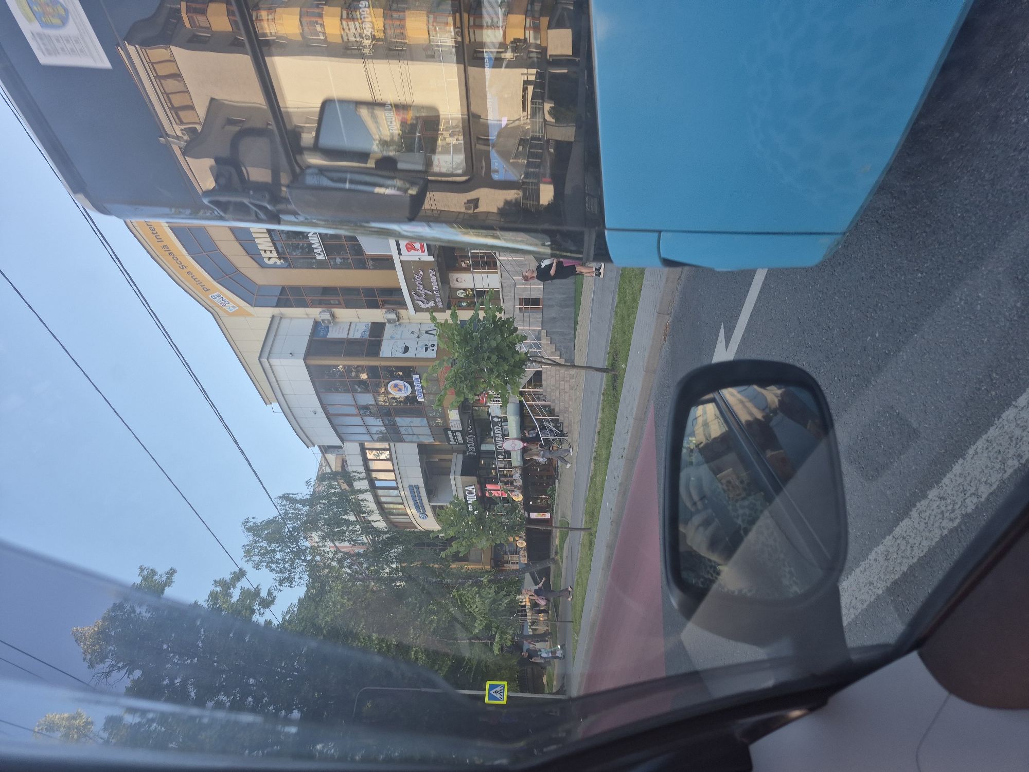 The image captures a street scene in Chișinău, Moldova, viewed from the interior of a vehicle. In the foreground, a blue bus is partially visible, occupying the right side of the frame. The bus's side and side mirror are prominent. The side mirror reflects a person holding a phone and wearing a leopard-print garment, along with some of the surrounding environment.
The street itself features a red-colored bike lane and a gray asphalt road with white lane markings. Several individuals are visible on the sidewalk and crossing the street. One person is on a skateboard.
On the left side of the image, a yellow pedestrian crossing sign with a blue symbol is mounted on a pole. Trees line the sidewalk, providing shade.
In the background, a multi-story building with large windows and several storefronts lines the street. Signs on the building include "SEMINARIUM KAMIN," "Prima Școală Internațională," "NICA," "Factory Outlet," and "LOMBARD." The architecture suggests a commercial and educational area. The sky is clear and blue, indicating daylight.