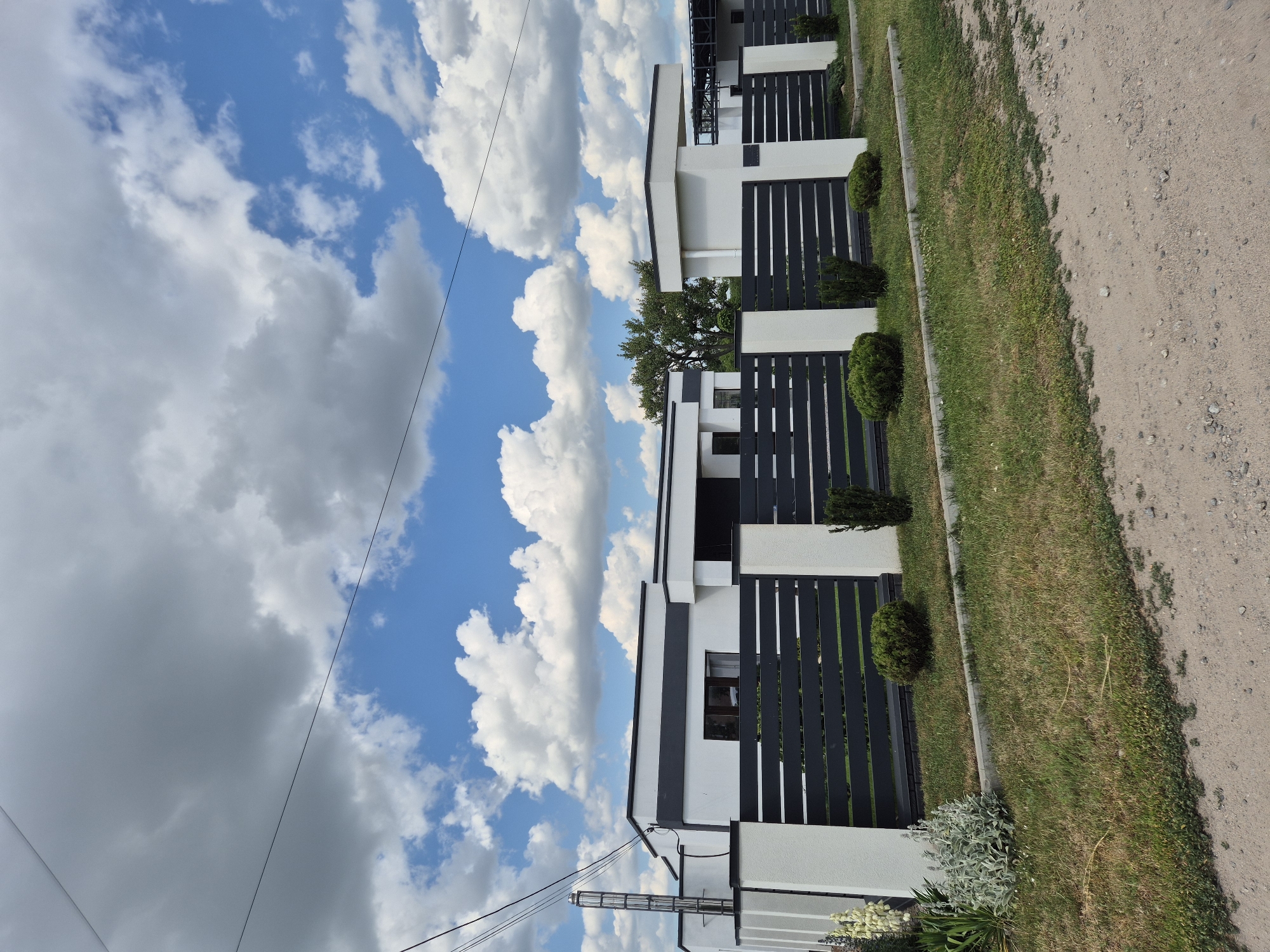 This is a sideways, eye-level, full shot of a modern white house with a dark gray fence and manicured greenery. The house has a clean, minimalist design with dark horizontal slats in the fence and windows. Small, rounded evergreen bushes are strategically placed along the fence line. The scene is bordered by a well-maintained lawn on the left and a gravel path on the right. The sky above is a vibrant blue, dotted with fluffy white and gray clouds, suggesting a bright, possibly windy, day. The overall impression is one of a well-kept, residential property in a suburban or rural setting. The orientation of the image suggests it was taken on a mobile device held vertically. No people are visible in the frame. There is no visible text.