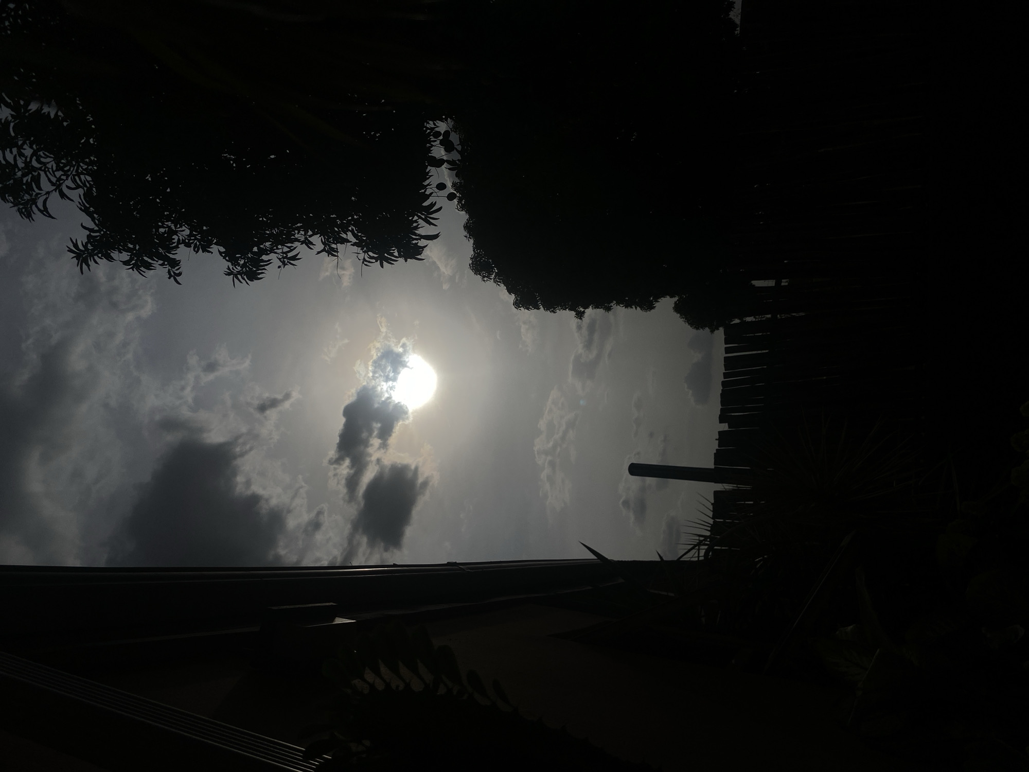 The image presents an upward view of a partially overcast sky, dominated by a bright, circular sun, partially obscured by clouds. The sky exhibits various shades of gray, with lighter tones around the sun and darker, more voluminous clouds scattered throughout.
Dark silhouettes of dense tree foliage or large plants frame the upper left and upper right sections of the image, contributing to a high-contrast aesthetic. Along the bottom edge, a dark, horizontal structure, possibly a roofline with visible ridging or corrugated elements, stretches across the frame. On the right side, extending upwards from this roofline, is a dark, slatted structure resembling a wooden fence or wall, next to which a slender, dark pole stands vertically.
No specific activity or event is taking place. The presence of the sun indicates it is daytime, with the dramatic lighting suggesting either morning or late afternoon. The scene captures the atmospheric conditions and the interplay of light and shadow, highlighting the natural environment and man-made structures from a low vantage point. The elements are consistent with an outdoor setting in a location like Barden Yana, Nigeria. There is no visible text in the image.