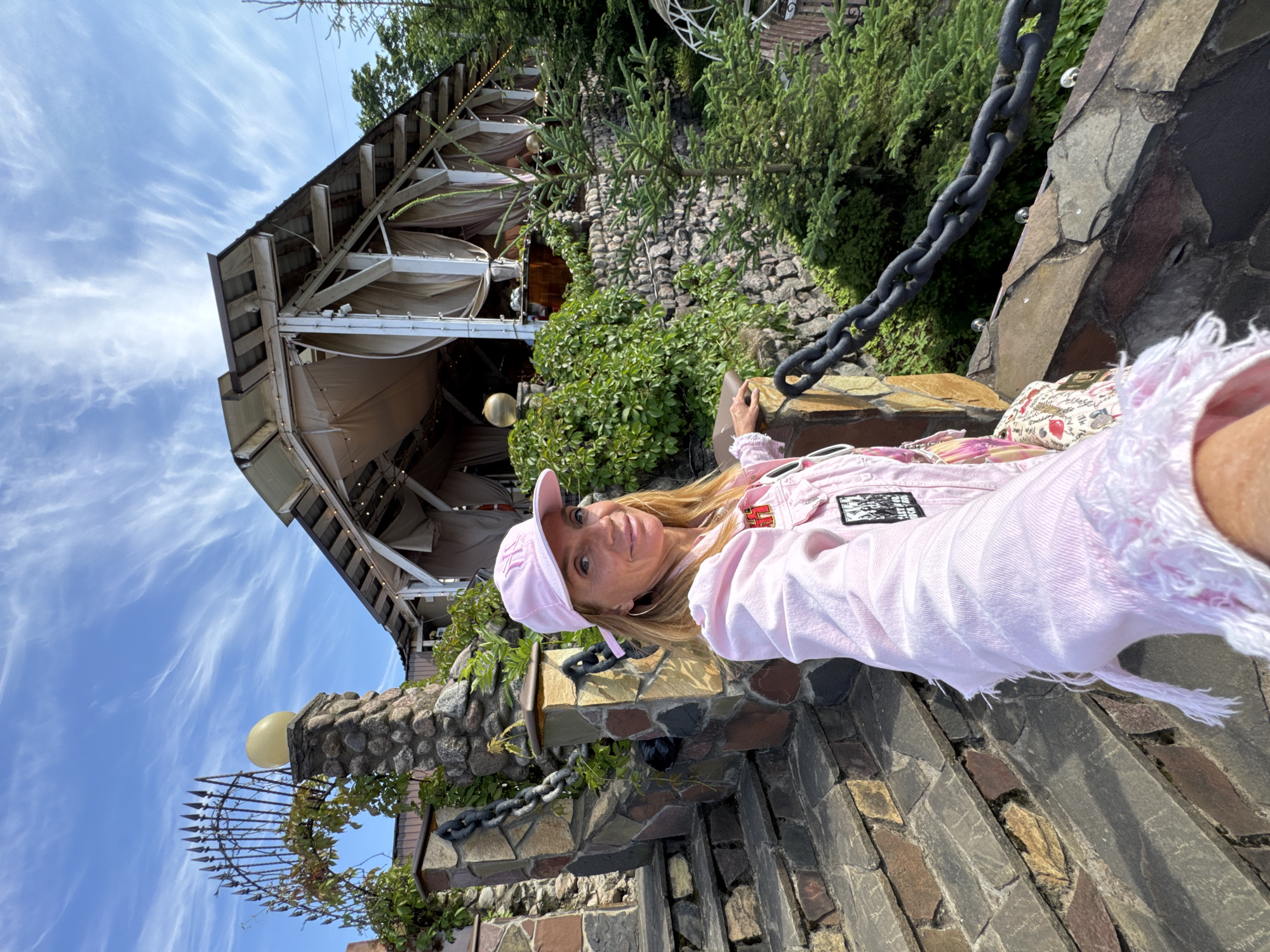 The image shows a woman in a pink baseball cap and a pink jacket with distressed fringes, leaning on a stone structure. She has blonde hair and is smiling at the camera. Behind her, a building with a rustic facade and a covered outdoor seating area is visible. The building is surrounded by lush greenery and tall evergreen trees. A large, dark metal chain runs along the stone structure in the foreground, and a decorative metal fence with pointed finials is also visible. The sky is blue with wispy white clouds. The scene appears to be set during the daytime, with bright sunlight casting shadows. The overall impression is one of a relaxed, outdoor setting, possibly a cafe or restaurant, in a natural environment. There is no visible text in the image.