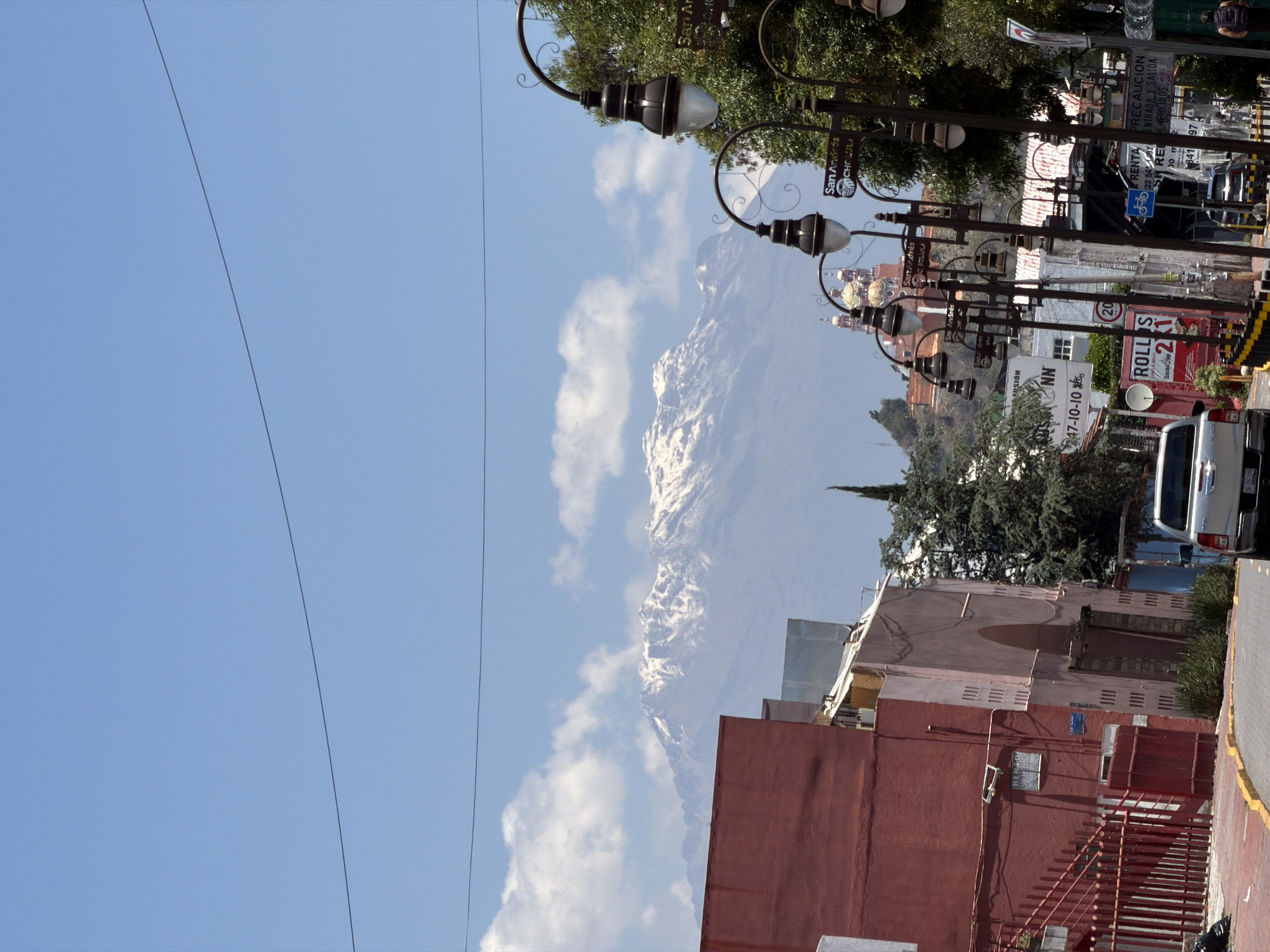 The image captures a view of San Andrés Cholula, Mexico, showing buildings and street elements with a mountainous backdrop. A deep blue sky occupies the left side of the frame, punctuated by electrical lines. In the distance, a snow-covered mountain is visible. Street lamps with decorative metalwork and signs reading "San Andrés Cholula" line the street, partially obscuring the mountain. Buildings with facades of red and tan hues are visible. A white van is parked near the right edge of the frame. Signage is present, including one advertising "Rollos 2x1". Trees and other greenery are interspersed among the buildings.