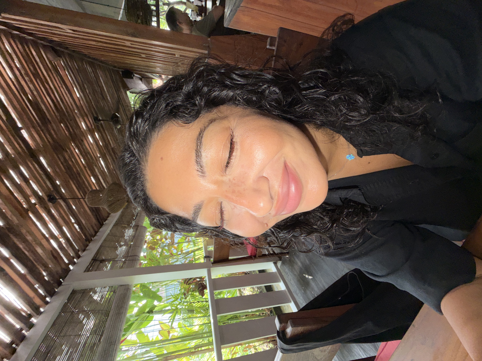 The image captures a serene moment featuring a woman in a semi-outdoor setting, consistent with the tropical environment of North Kuta, Indonesia. The photo is oriented with the woman's head on the left, looking upwards with her eyes gently closed and a soft, contented smile on her lips. She has dark, curly hair and appears to be wearing a black top or dress. A delicate silver necklace with a small, light blue, possibly butterfly-shaped, pendant is visible around her neck. Light freckles are visible across her nose and cheeks.
In the background, a rustic wooden ceiling composed of parallel slats creates striking patterns of light and shadow as bright daylight filters through. Below this, white painted wooden railings suggest an open-air balcony or patio area. Beyond the structure, lush green tropical foliage is abundant, contributing to the natural and peaceful atmosphere. A woven pendant light fixture hangs from the wooden ceiling. Another individual, partially obscured and possibly a man wearing a green shirt, is visible in the upper background, seated at a wooden table.
The strong, natural lighting indicates it is daytime, likely a sunny day. The scene evokes a feeling of relaxation and tranquility, suggesting the woman is enjoying a calm moment in a beautiful, natural environment, possibly at a resort, cafe, or restaurant. No text is visible in the image.