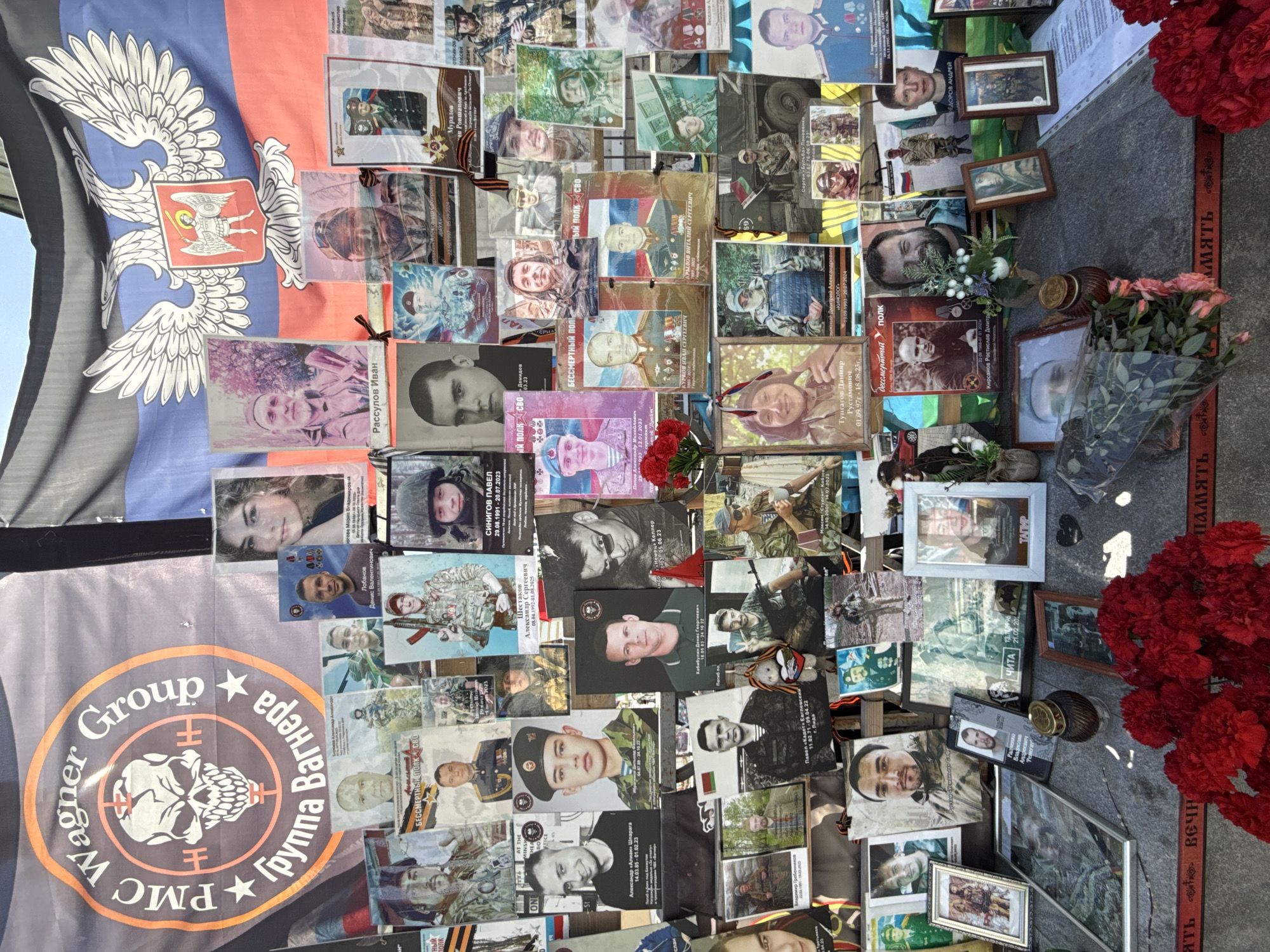 The image depicts a memorial, likely located in Moscow, Russia, based on context clues and the Cyrillic alphabet text. The memorial consists of a display of numerous photographs and memorial cards of individuals, predominantly men, but with at least one identified female in the upper left. Several photos depict individuals in military or paramilitary attire.
A flag bearing the emblem of the self-proclaimed Donetsk People's Republic (DPR) is visible in the upper left. Another flag displaying the logo of the Wagner Group, a Russian private military company, is in the lower left corner. Both flags are displayed beside the photographs.
The photos are arranged on a stone surface, possibly a gravestone or memorial slab. Fresh red carnations and other flowers, along with framed photographs and candles, are arranged at the base of the photo display. A red ribbon with the Cyrillic inscription "ВЕЧНАЯ ПАМЯТЬ" (Eternal Memory) borders the stone surface.