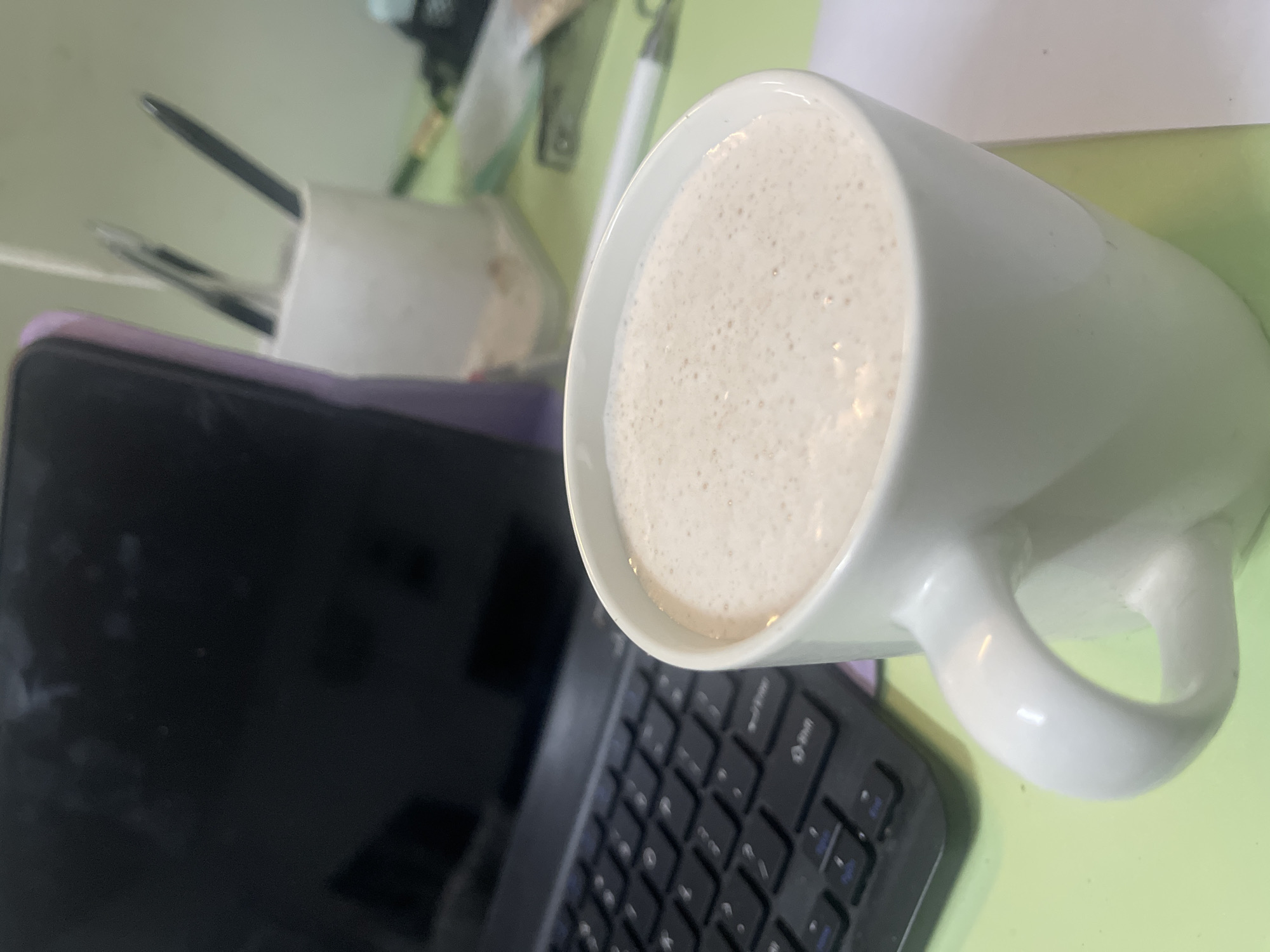 The scene shows a close-up, top-down view of a workspace with a white mug filled with a light-colored beverage, possibly a latte or cappuccino, occupying the right side of the frame. A black tablet and a wireless keyboard are situated to the left and slightly below the mug. Behind the tablet, there is a white container holding pens or pencils. The background appears to be a light green surface, potentially a desk or tabletop. The setting is likely an office or home workspace, but the precise location in the Chongqing High-tech Zone, China cannot be determined from the image. There are no people visible in the photograph.
