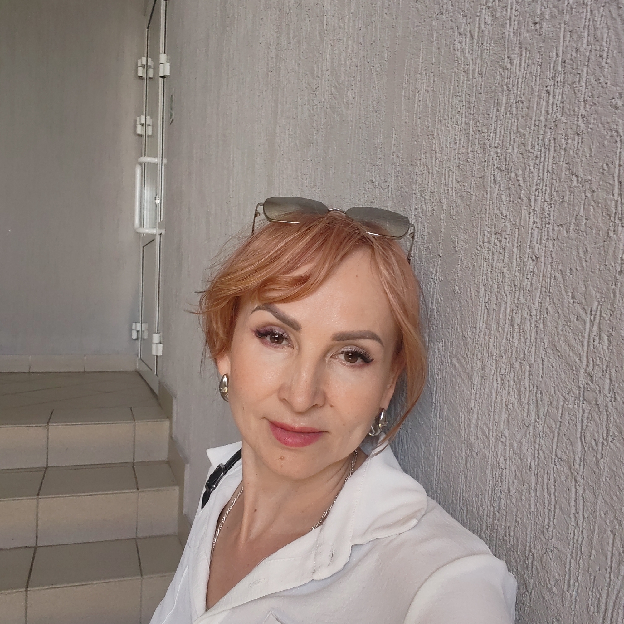 A middle-aged woman with short, reddish-blonde hair and sunglasses resting on her head stands close to a rough, grey wall. She wears a white collared shirt, silver hoop earrings, and a silver necklace. A black strap is visible near her left shoulder. In the background, there are light grey stairs leading to a white door with metal hardware. The location appears to be in Chaikovsky, Russia.