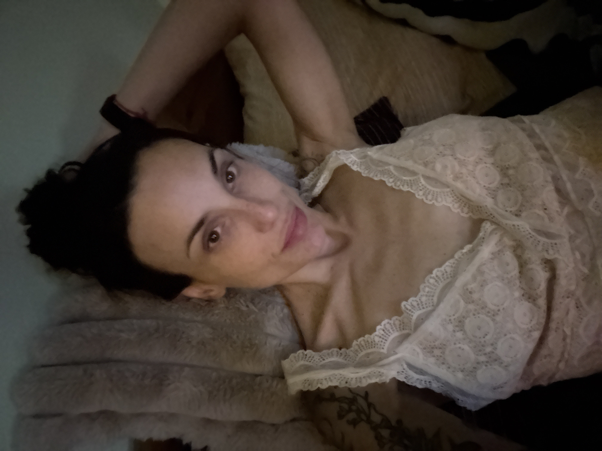 A woman is lying on a light brown furry pillow, facing the camera. She has dark hair pulled back in a bun, fair skin, and brown eyes. She is wearing a white lace top. Her right arm is bent behind her head and she has a black bracelet on her right wrist. Her left arm is visible resting on the pillows. A portion of her arm has a tattoo. A light brown pillow is behind her head and a black striped cushion is in the background. The location is unknown, but has not been identified as Pleven, Bulgaria.