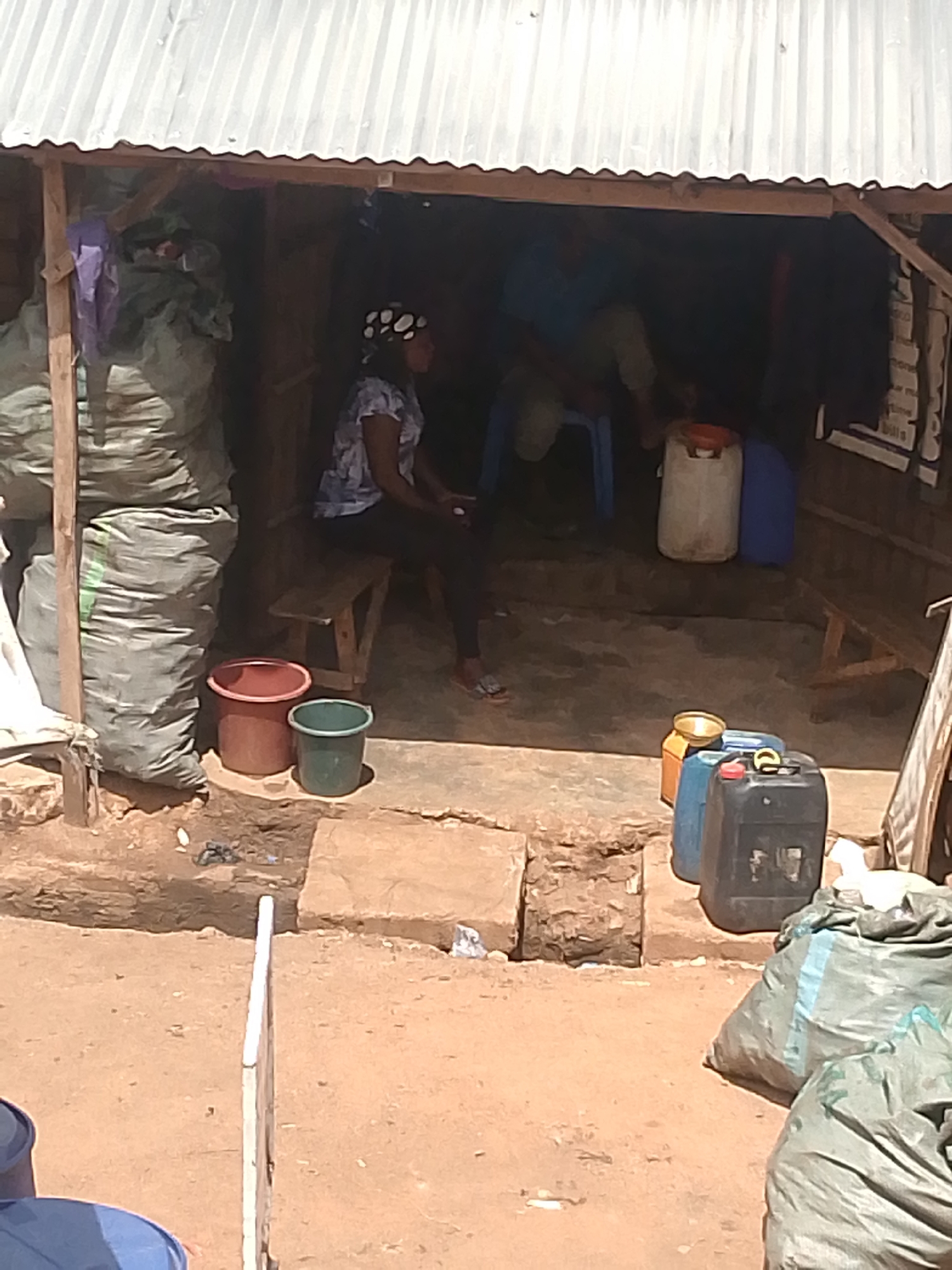 The image shows a scene in Bukuru, Nigeria. Under a corrugated metal roof supported by wooden beams, a woman with a spotted head covering sits on a wooden bench. Inside, a man sits on a blue plastic chair. Next to the woman are a red and a green plastic bucket. Inside are a white and a blue plastic container. To the right are an assortment of yellow, blue, and black plastic containers. Bags of unidentified materials are stacked on the left and right sides of the open-fronted structure. The ground is dirt with concrete patches.