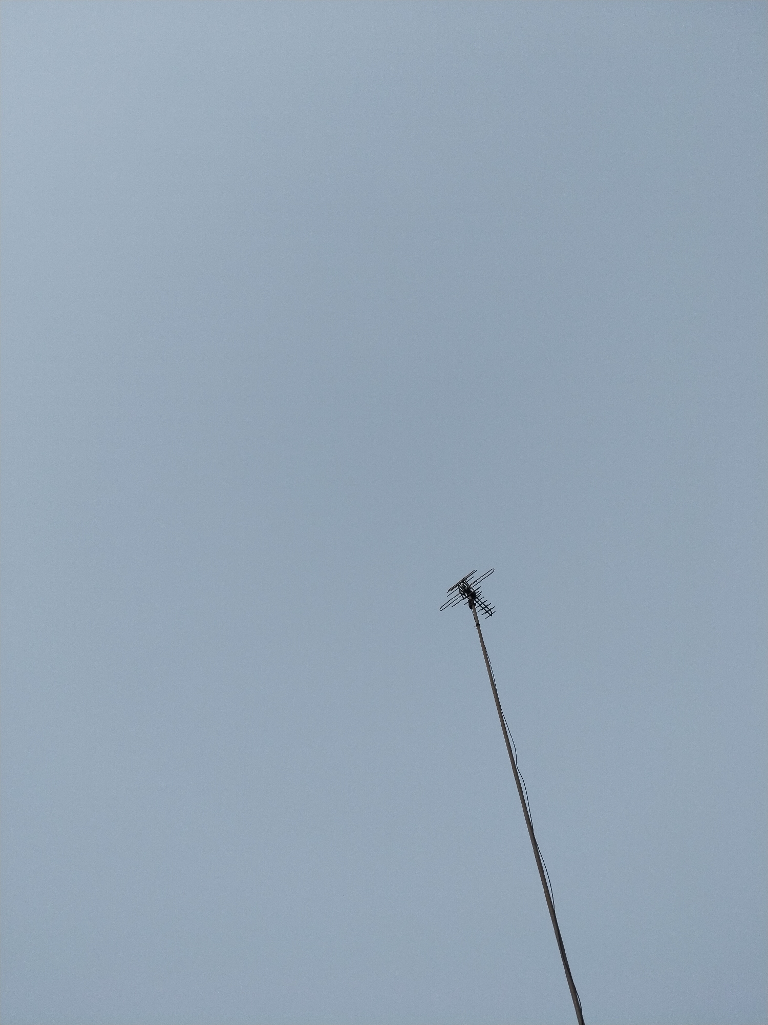 The image shows a close-up, low-angle view of a television antenna mounted on a pole against a clear, pale blue sky. The antenna is a traditional directional type with multiple elements arranged in a cross pattern at the top, and it is attached to a dark, slender pole that extends diagonally from the bottom right corner of the frame upwards towards the antenna. A dark cable runs down the pole. The background is uniformly overcast sky, with no discernible landmarks or environmental features visible. The scene suggests an outdoor setting, likely a residential or commercial area where rooftop antennas are still in use. The specific location in Kampung Baru, Indonesia, cannot be determined from the visual information presented in the image itself.