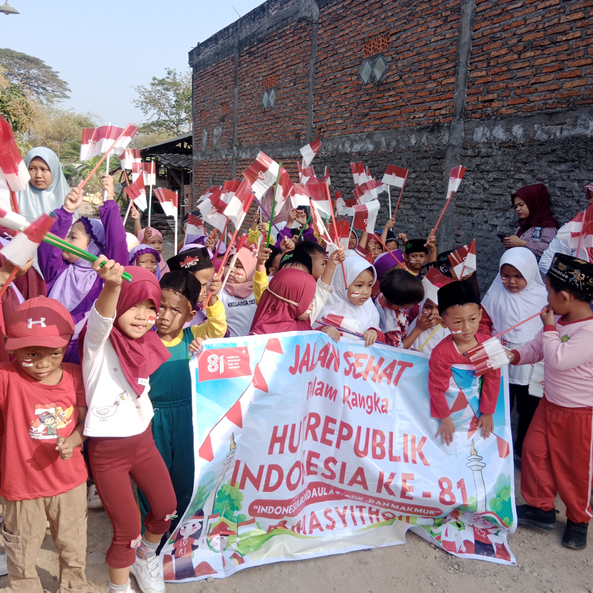 A group of young children and adults are gathered in what appears to be a street in Tegowanu, Indonesia, celebrating Indonesia's 81st Independence Day. Many of the children are holding small Indonesian flags. A large banner in the foreground reads "JALAN SEHAT DALAM RANGKA HUT REPUBLIK INDONESIA KE-81". The background consists of a brick building and some trees. The children are dressed in a variety of clothing, including red shirts, purple shirts, and headscarves. Some of the children have face paint.