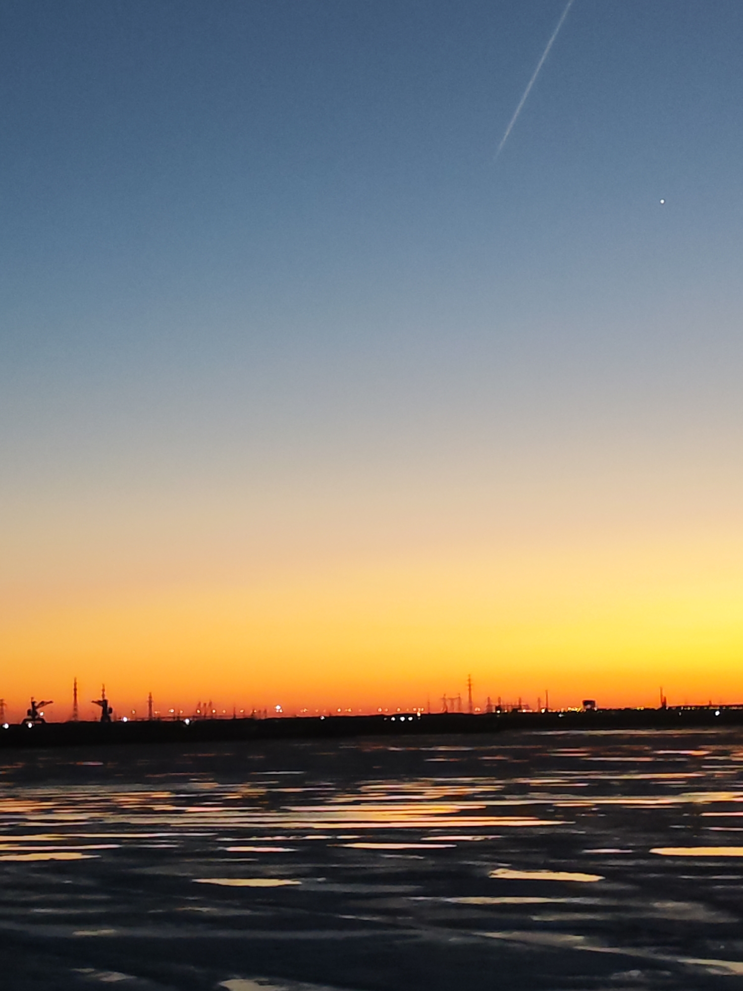 The image depicts a twilight scene in Chaikovsky, Russia. The sky transitions from blue to orange, with a contrail and a bright celestial object visible. Below the horizon, a silhouette of industrial infrastructure is discernible. The foreground features a body of water or a wet surface reflecting the twilight hues.