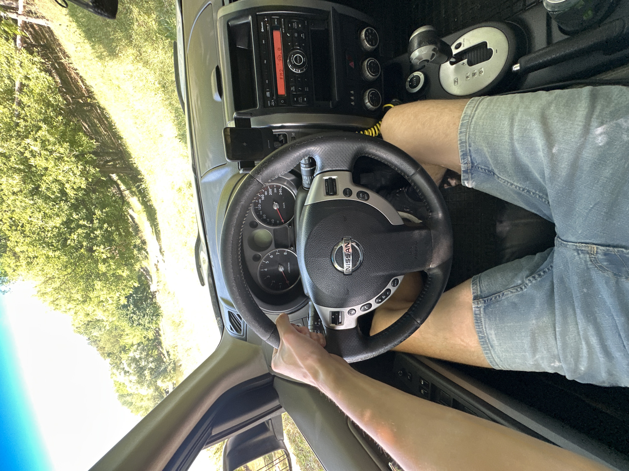 The image shows a first-person perspective from inside a Nissan vehicle, with the driver's left hand on the steering wheel. The driver is wearing denim shorts. Outside the car, lush green trees and a bright blue sky are visible, suggesting an outdoor setting. The car's dashboard is also visible, featuring the steering wheel with the Nissan logo, the instrument cluster, and the car's radio. The scene appears to be during daylight hours, likely in a rural or suburban area given the natural scenery. There is no specific indication of the location being Makiv, Ukraine.