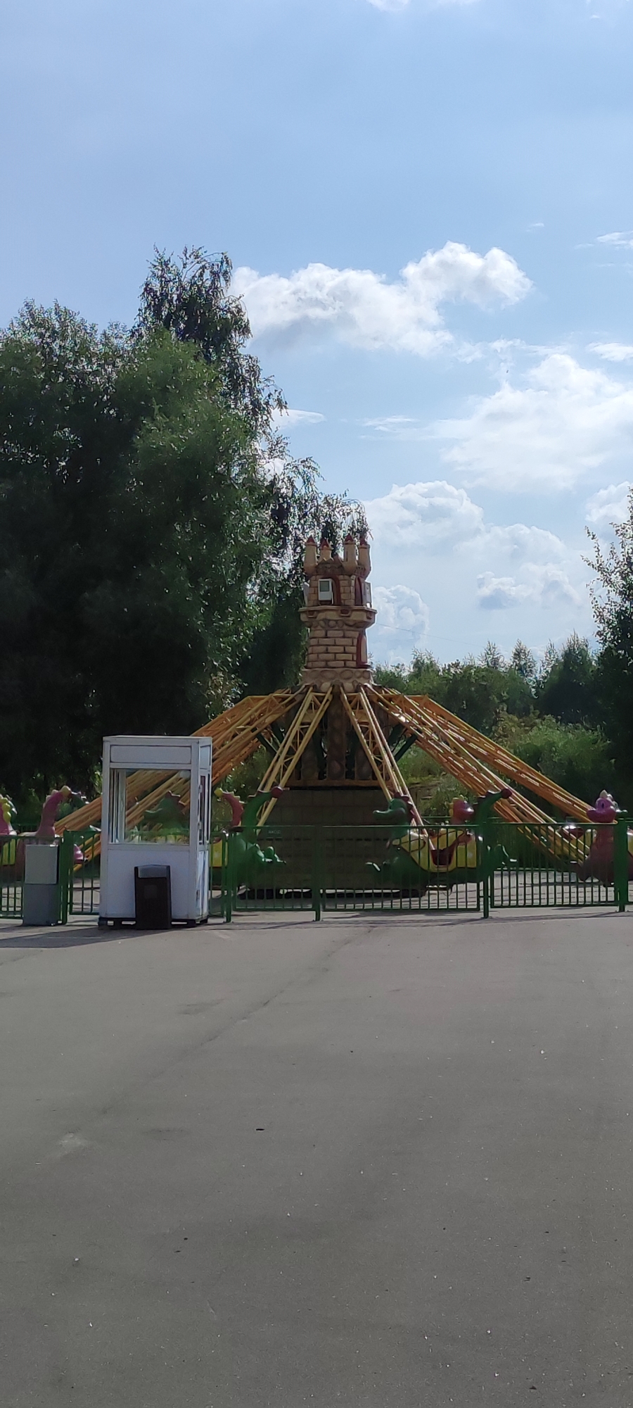 The image shows an amusement park ride with a castle-like structure at its center. The ride features several colorful dragon-shaped cars. A small, white ticket booth or control booth is visible to the left of the ride, with a black trash bin next to it. The ride is enclosed by a green fence. In the background, lush green trees and a blue sky with scattered white clouds are visible. The ground in front of the ride is paved with asphalt. The scene appears to be a bright, sunny day. This amusement park is located in the city of Khimki, Russia.