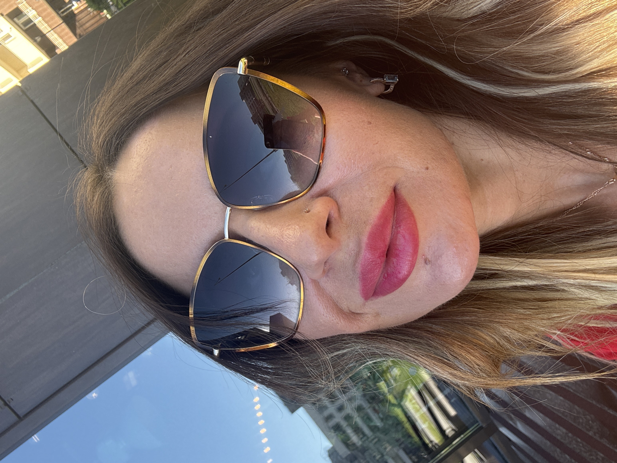 This media file is a close-up, angled selfie of a woman wearing large, gold-rimmed sunglasses with dark lenses. Her lips are painted a vibrant pink. She has long, wavy brown hair with blonde highlights. The background is blurred, but appears to be an outdoor setting, possibly a cafe or restaurant, with reflections of buildings and a blue sky visible in a reflective surface, likely a table or window. The lighting suggests it is daytime, and the overall mood appears relaxed and perhaps candid. No visible text is present in the image.