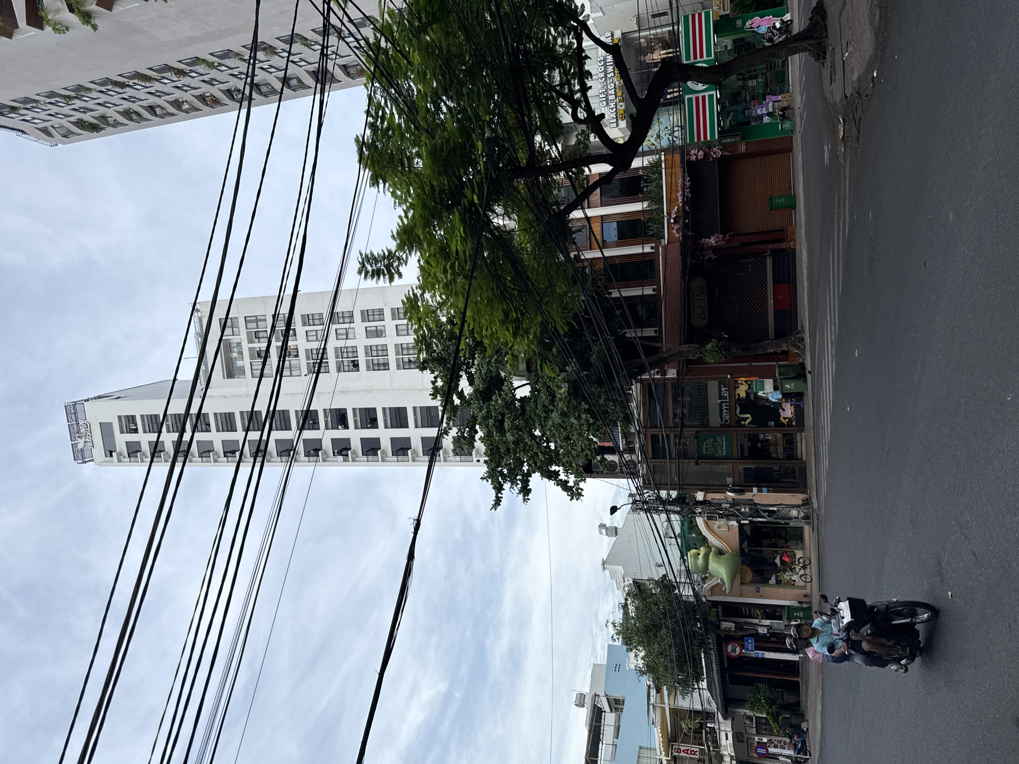 The image captures a street scene in Nha Trang, Vietnam, dominated by a tall, white, modern apartment building on the left. A dense network of black electrical wires crisscrosses the frame in the foreground, obscuring parts of the sky and the buildings behind them. A large green tree with lush foliage is positioned in the mid-ground to the right, its branches extending across the wires.
Below the tree, a row of shops and businesses line the street. One shop has a prominent green, cartoonish mascot figure above its entrance. Another shop, to the right, appears to be a 7-Eleven convenience store, identifiable by its red and green signage. A motorcycle carrying two individuals, a man driving and a woman passenger, is visible in the lower left quadrant of the image, moving across the asphalt road. The road is wet, suggesting recent rain, and there are white pedestrian crosswalk markings. The sky is overcast with a pale, diffused light. In the background, another tall white building, similar in style to the one on the left, is partially visible. The overall impression is of a typical urban environment in a tropical city, with a mix of modern architecture, dense infrastructure, and everyday street life.