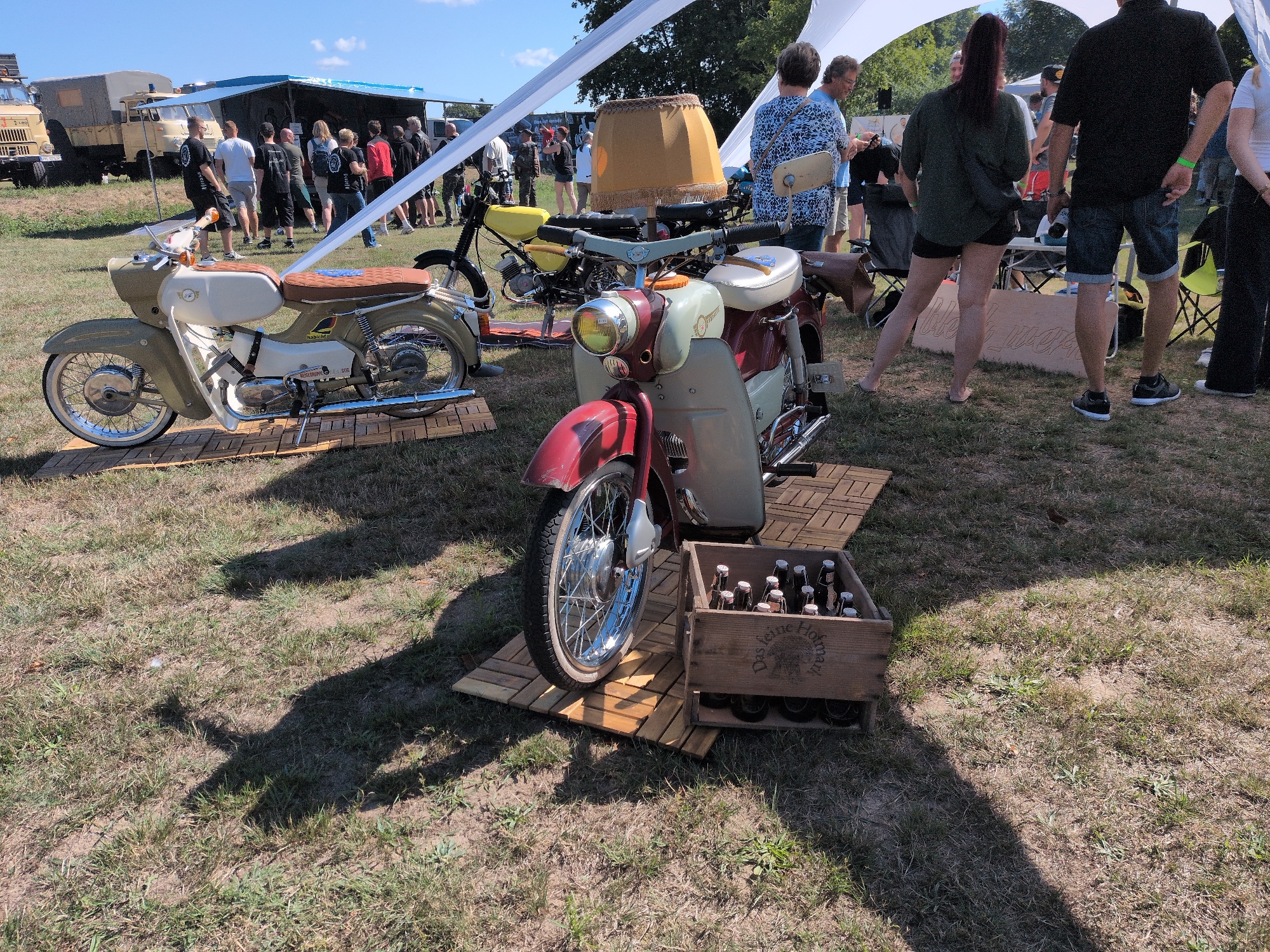 On a sunny day in Hohenferchesar, Germany, two vintage mopeds are displayed on a grassy area. The moped in the foreground is red and cream-colored, with a chrome front wheel and handlebars. It is parked on a wooden pallet. To the left and behind it, a second moped, olive green and cream, also sits on a pallet. A wooden crate filled with glass bottles, possibly beer, is positioned to the right of the foreground moped's front wheel. In the background, several people are gathered under a white tent, with two military-style trucks parked behind them. The sky is clear and blue.