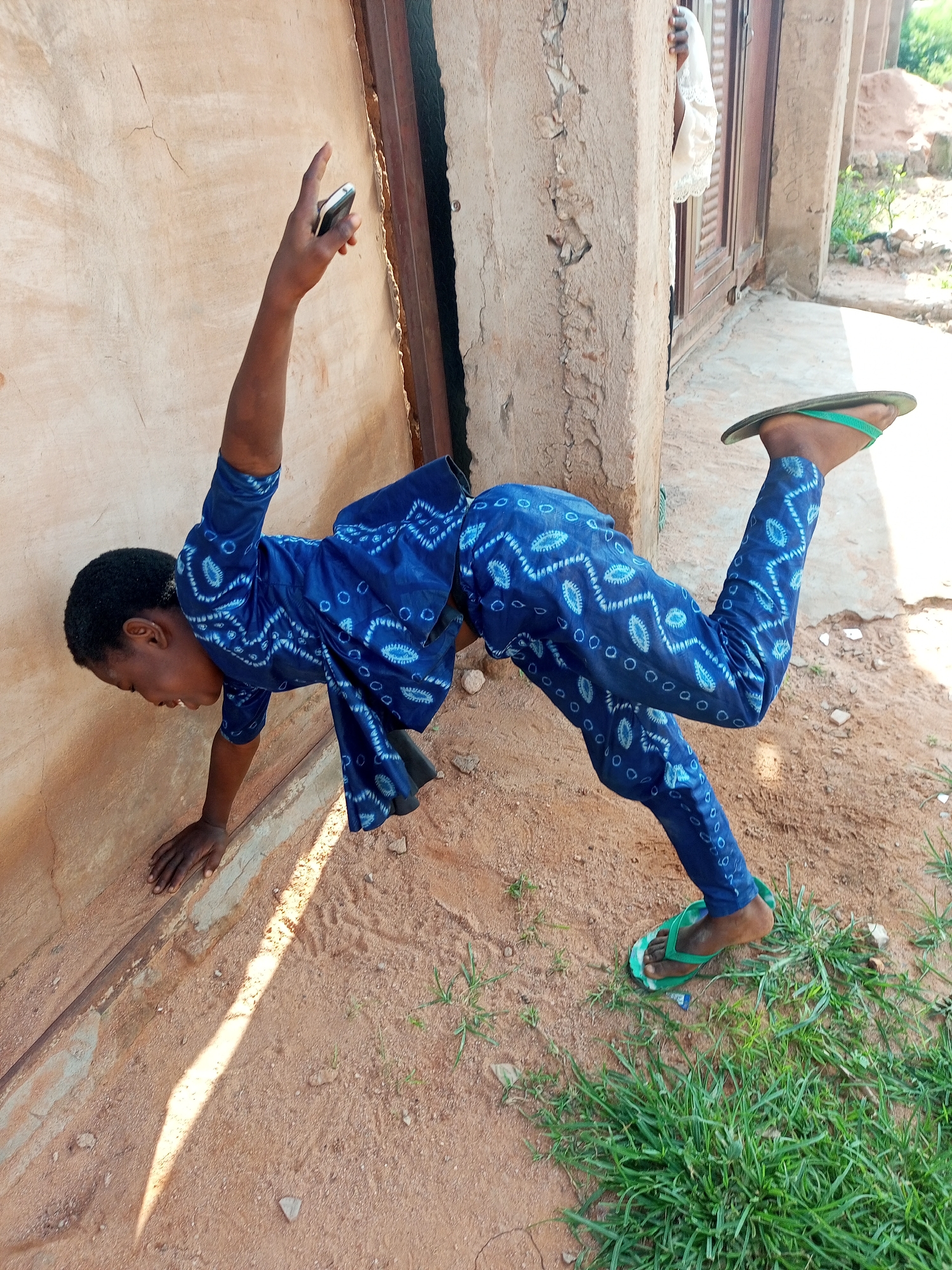 A person is performing a handstand against a textured, light-colored wall. They are wearing a blue patterned outfit and green flip-flops. The ground is sandy with patches of grass. In the background, a wooden door and a white piece of fabric are visible. The scene appears to be outdoors, possibly in a rural or less developed urban area, consistent with parts of Tempure, Nigeria. The lighting suggests it is daytime.