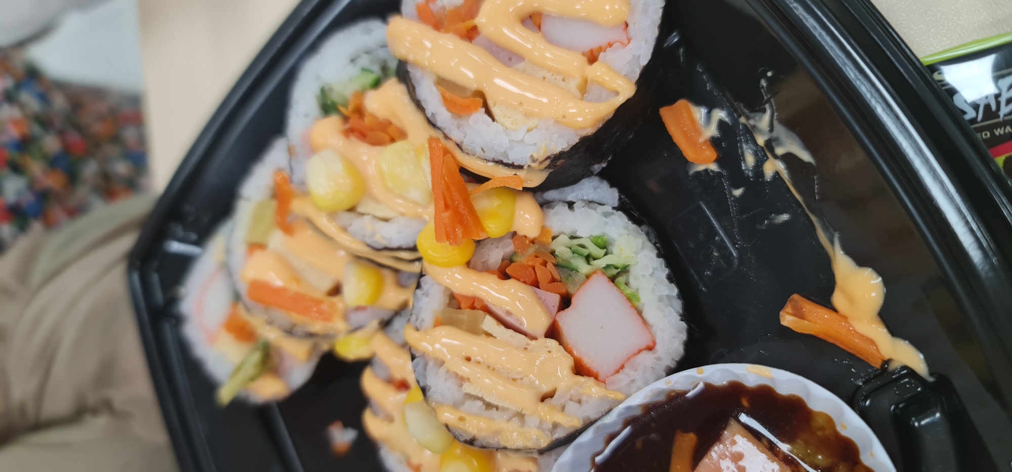 A close-up, slightly angled shot captures multiple pieces of sushi or kimbap arranged in a black plastic take-out tray. The sushi rolls feature white rice on the exterior, wrapped around a dark seaweed layer, and filled with various ingredients including light orange crab sticks (surimi), shredded green vegetables (possibly cucumber), shredded orange carrots, and bright yellow corn kernels. A creamy, light orange sauce is generously drizzled over the top of the rolls, and some drips are visible in the tray.
In the bottom right corner, a small, white plastic cup filled with a dark brown dipping sauce (likely soy sauce) is partially visible. The background is blurred, showing a warm-toned fabric object (possibly clothing) in the lower left, and a colorful, patterned object further back. To the upper right, a black and green item with partially visible text, including "SAB" and "ED WA", can be discerned. The overall impression is a prepared meal or snack, likely consumed in an indoor setting in Đà Nẵng, Vietnam.