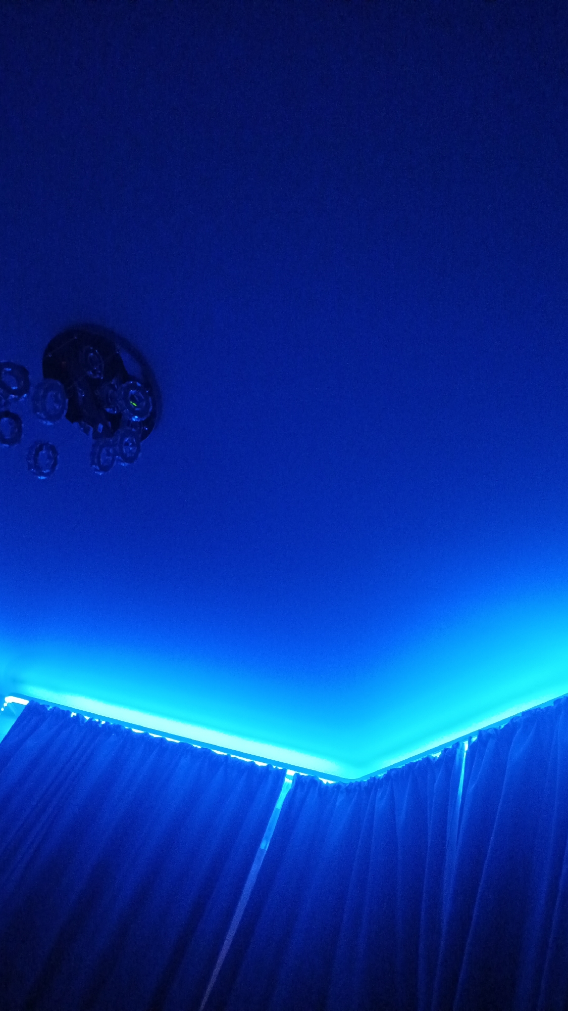 A ceiling fan with attached crystal or glass embellishments is visible in the upper left corner of the image. The fan and embellishments are dark in color. The ceiling is illuminated with a vibrant blue light, creating a gradient effect that is brightest along the edge of the ceiling and fades upwards. The lower portion of the image shows dark blue curtains, likely hanging from the ceiling. The setting appears to be an interior space. There is no discernible information about Zvenigorod, Russia.