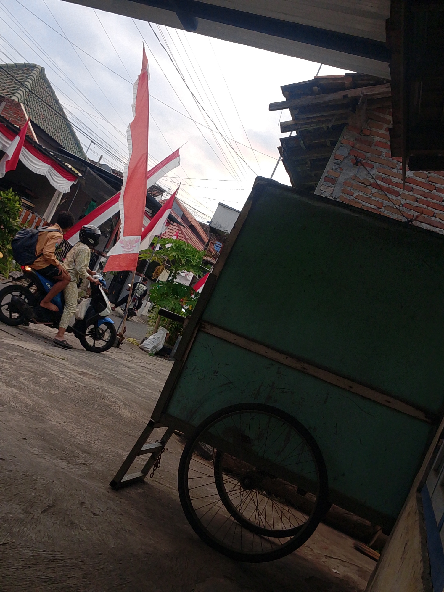 In an urban environment, possibly in Banyuwangi, Indonesia, a scene unfolds beneath an overcast sky. The foreground is dominated by a large, weathered green cart with a wooden frame, its two spoked wheels partially visible. The cart is positioned on a paved surface that shows signs of wear.
In the midground, two individuals are riding a blue motorcycle. The rider, wearing a helmet, is dressed in light-colored patterned clothing. The passenger, seated behind the rider, wears a tan jacket and has a dark backpack slung over their shoulder. Their feet are bare. The motorcycle appears to be moving.
The background features a row of buildings, some with tiled roofs, adorned with red and white banners, suggesting a local celebration or event. Power lines are strung across the sky, indicating an urban setting. The overall impression is one of everyday life taking place amidst festive decorations.