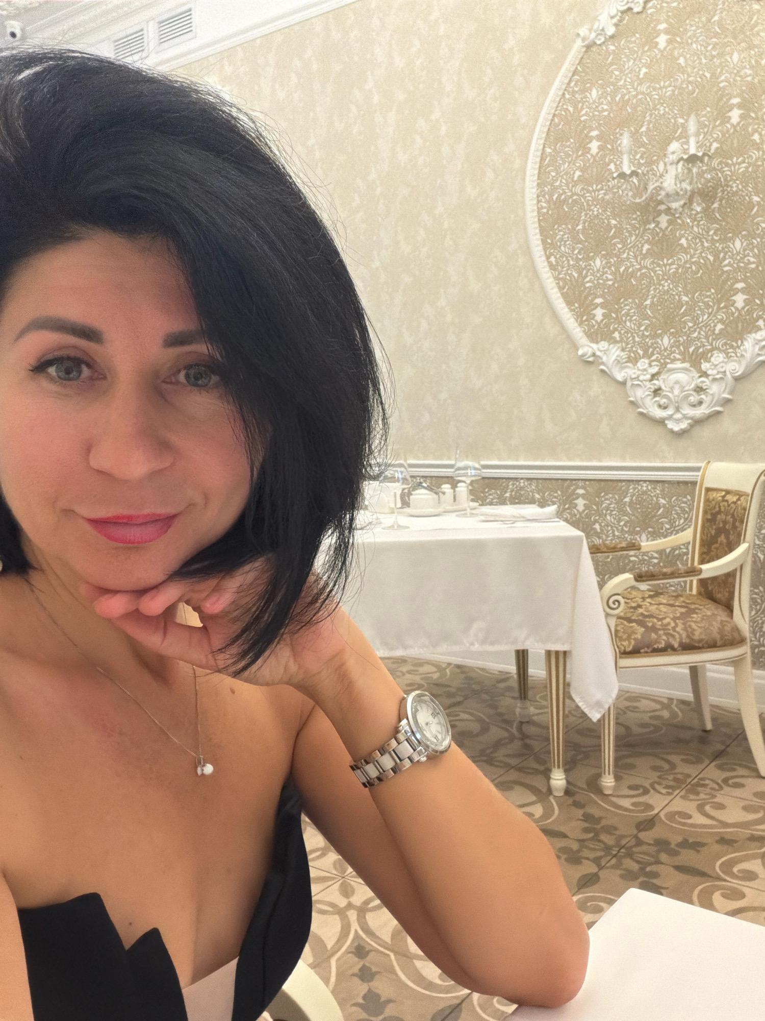 The image is a selfie taken by a woman with dark hair and blue eyes, wearing a black strapless dress and a watch on her left wrist. She is indoors, in what appears to be a restaurant or dining area, with a table set for a meal in the background. The walls are decorated with ornate wallpaper and a decorative wall sconce. A chair with a patterned cushion is visible next to the table. The floor has a patterned tile design. The woman is resting her chin on her hand and looking at the camera with a slight smile. There is no information or visual cue in the image to suggest the location is the city of Angarsk, Russia.