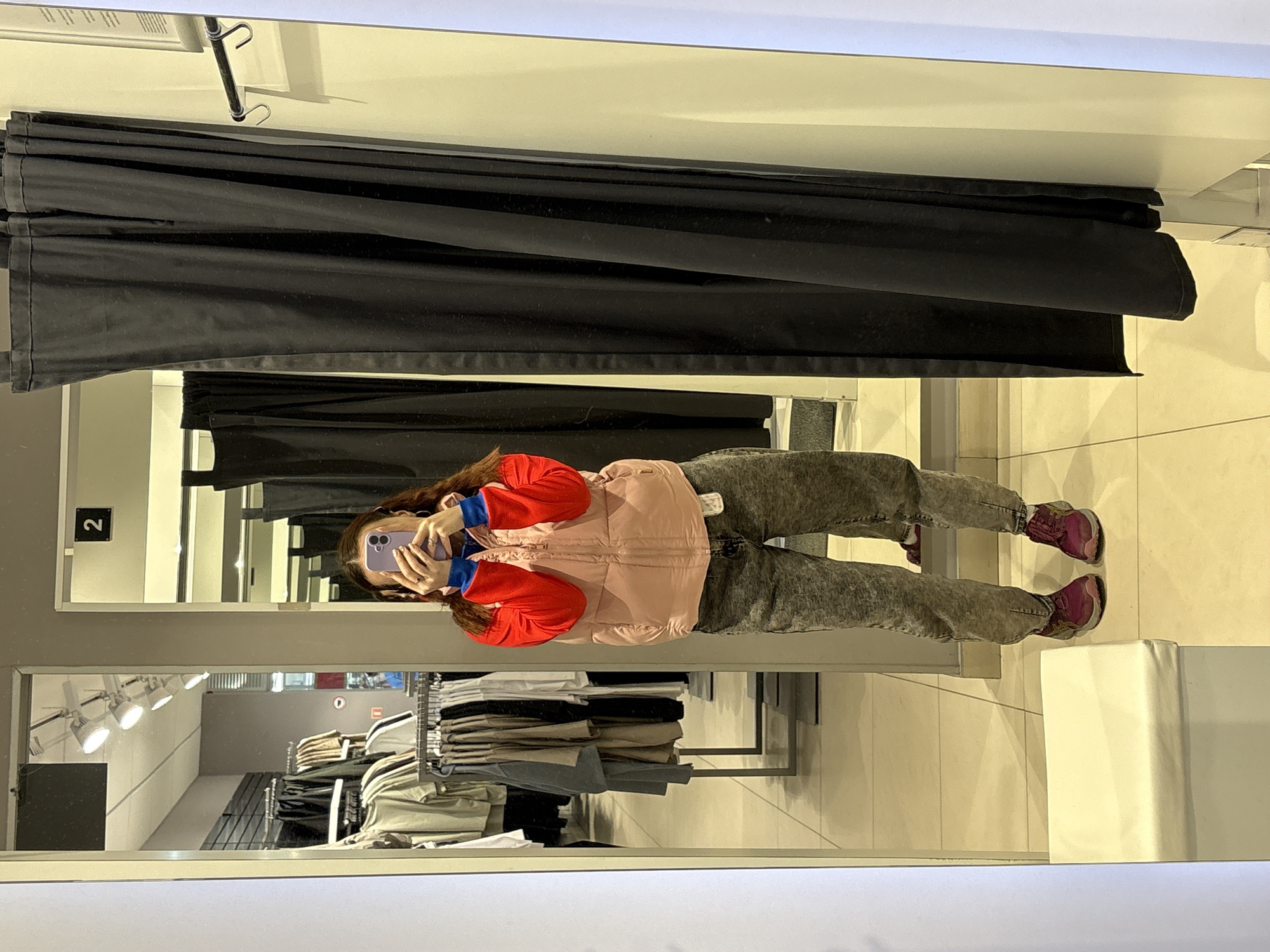 The image shows a person taking a photo of themself in a fitting room. The individual is wearing a light pink puffer vest over a red and blue long-sleeved shirt, grey jeans, and purple sneakers. The person is holding a purple smartphone in their hands, presumably to capture the image in the mirror. The fitting room has a black curtain as a backdrop, and another fitting room is visible through the mirror. Clothes racks with various garments are visible in the reflection, suggesting a clothing store setting. The location is a fitting room in an unidentified store, city, and state.