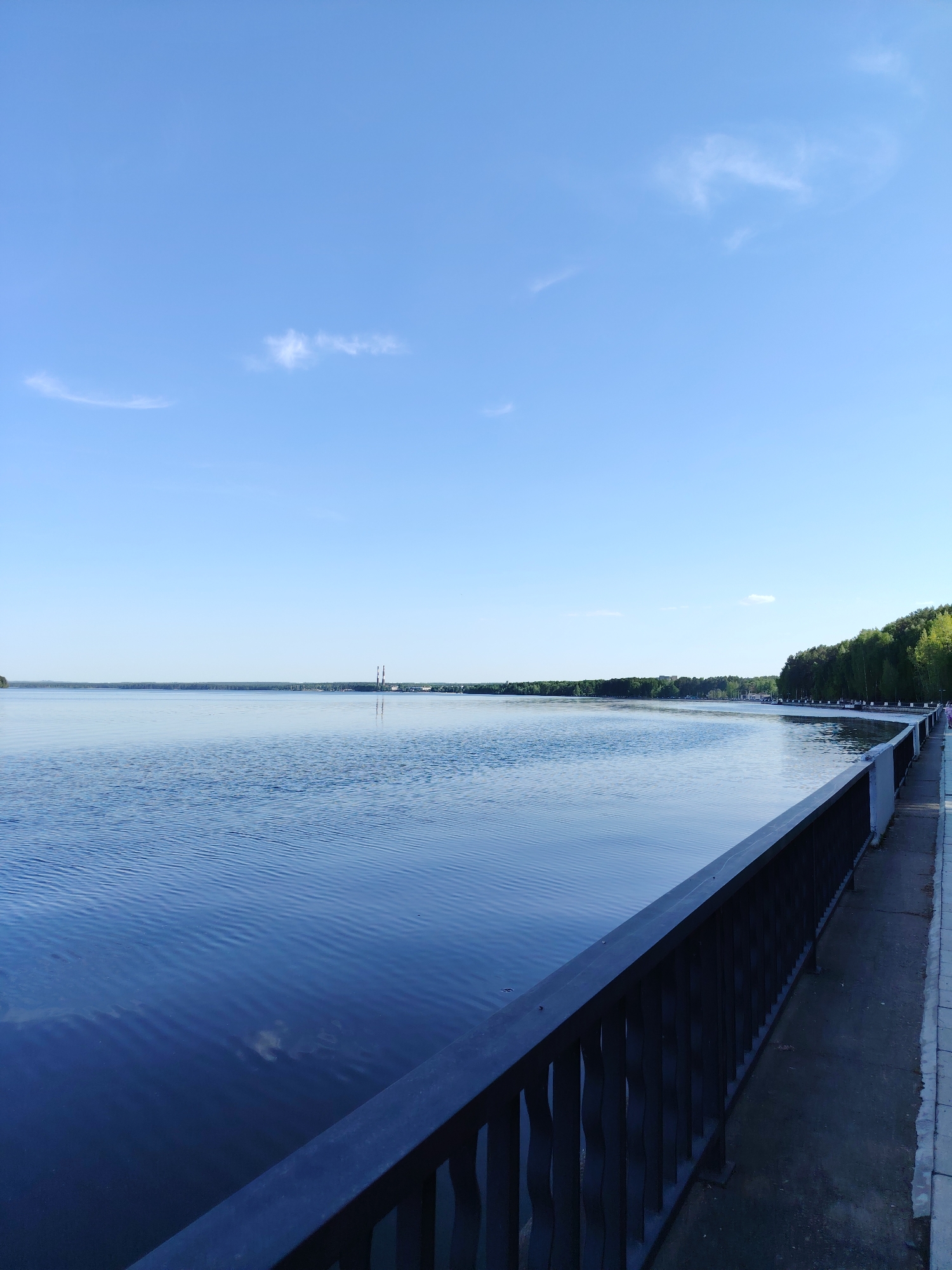 The image shows a view of the Votkinsk Reservoir in the city of Chaikovsky, Perm Krai, Russia. The body of water is calm, with slight ripples on the surface. A metal railing runs along the bottom right of the frame, separating a paved walkway from the water's edge. In the distance, the opposite shore is visible with a line of trees and some industrial structures with tall chimneys. The sky is mostly clear with light blue hue and a few scattered clouds. The lighting suggests it is daytime, likely in the afternoon or early morning. The general mood is peaceful and serene.