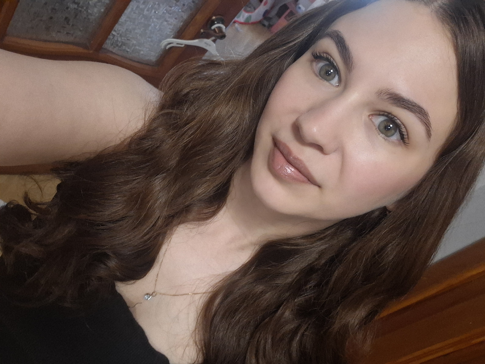 A close-up, selfie-style photograph captures a woman with light skin, brown hair, and greenish-hazel eyes. She is facing the camera at a slightly upward angle. Her eyebrows are well-defined, and she wears lip gloss. A delicate gold necklace is visible around her neck. The background includes a wooden window frame and what appears to be part of a room, indistinctly seen.
There is no information to verify the location as Chaikovsky, Russia, in the image.