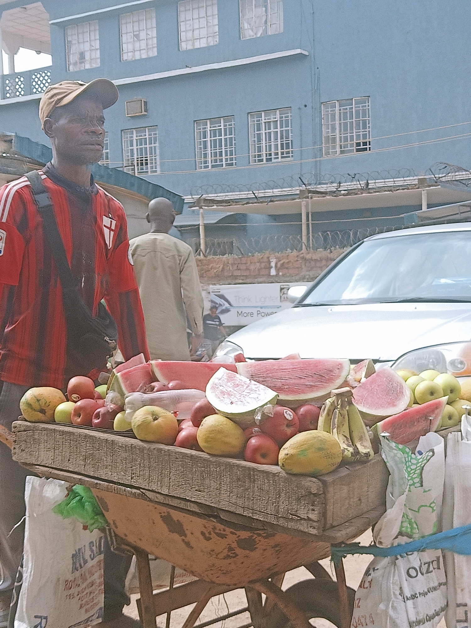 A fruit vendor in Jos, Nigeria, stands with a wooden cart laden with watermelons, apples, bananas, and other fruits. He is wearing a red and black striped shirt with a baseball cap and a bag slung across his shoulder. Behind him is a light-colored building with many windows and a vehicle. Another man is walking behind the cart and vendor. The street scene is busy. A fence topped with barbed wire is visible in the background.
