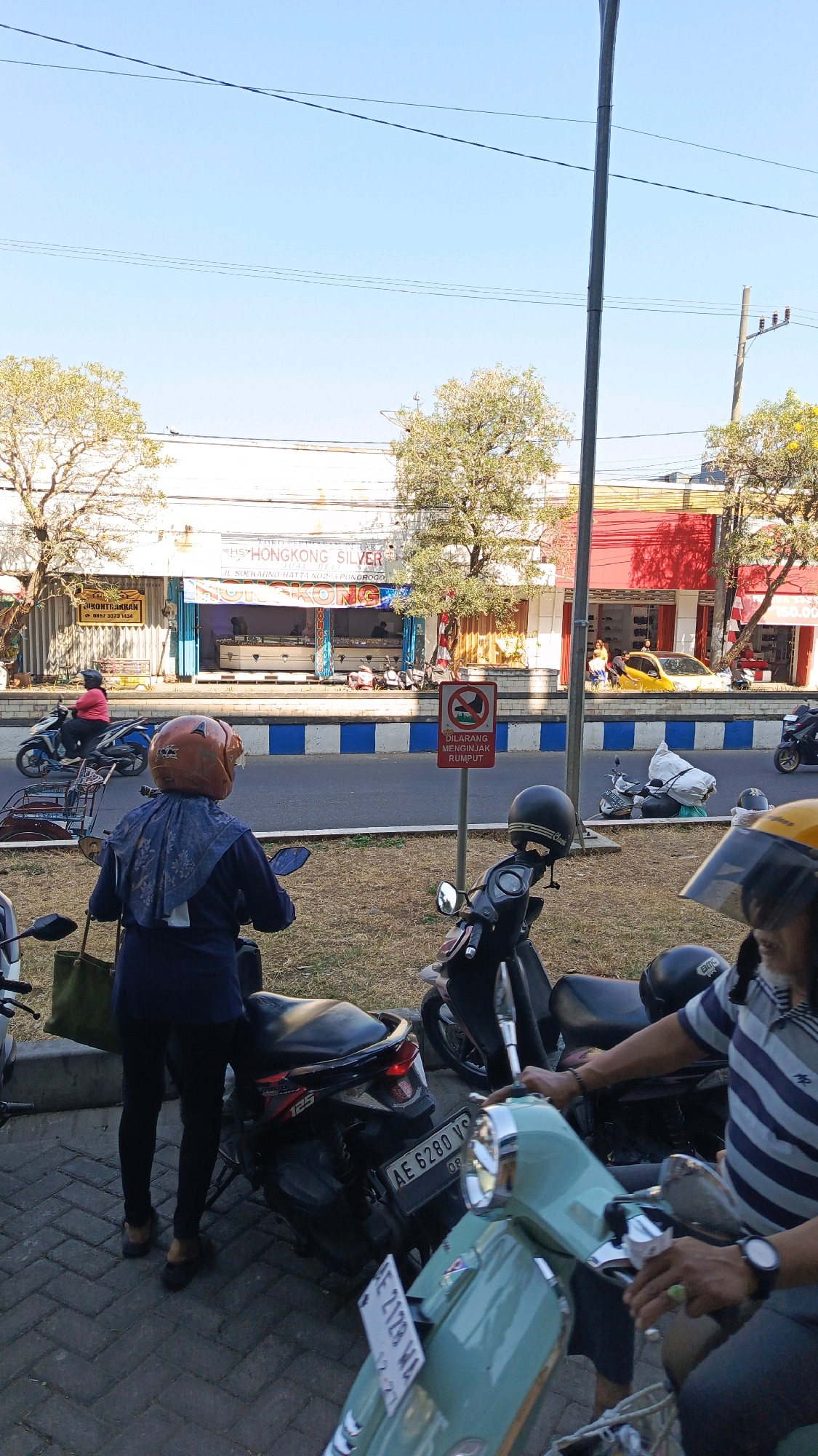 The image depicts a street scene in Ponorogo, Indonesia, likely on a sunny day. In the foreground, several motorcycles are parked or in use. A person wearing a blue jacket, black pants, and a brown helmet is on a dark blue motorcycle, holding a green bag and looking down at their phone. To the right, another person wearing a blue and white striped shirt and a black helmet is on a light green scooter. Further back, a yellow car is parked on the side of the road, and several other motorcycles are visible.
The background shows a row of buildings, including a shop labeled "HONGKONG SILVER" which sells jewelry. There is a red building to the right of the silver shop. A street with traffic runs in front of the shops. A red sign on a pole near the street reads "DILARANG MENGINJAK RUMPUT" which translates to "DO NOT STEP ON GRASS." Trees with green and yellow leaves line the street. Power lines are visible across the clear blue sky. The ground in the foreground is paved with interlocking bricks.