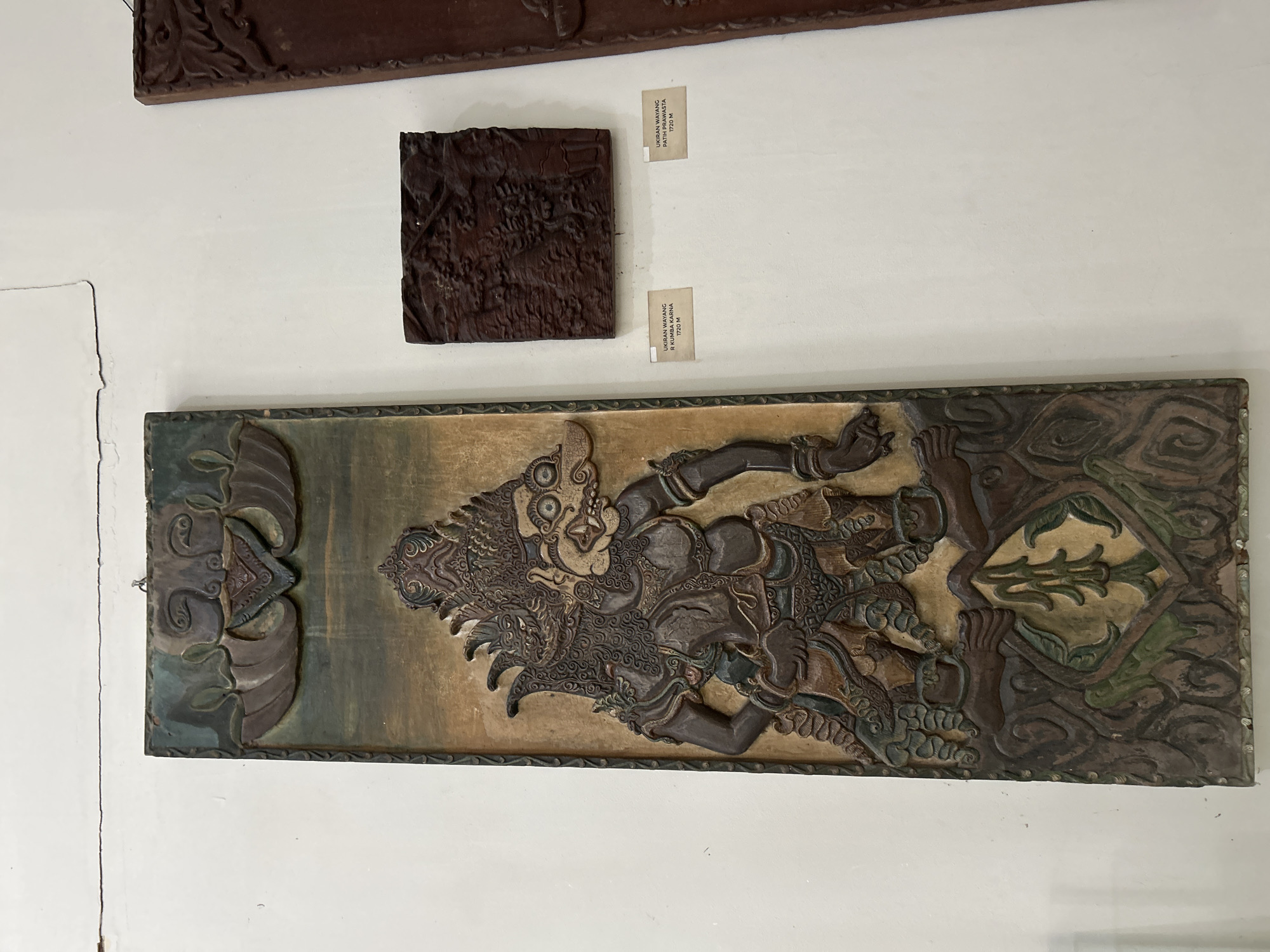 This media file captures a display of carved artworks on a white wall, likely in a museum or gallery setting in Cirebon, Indonesia. The primary focus is on two dark wooden relief carvings.
The larger, lower carving is a horizontal panel featuring a dynamic, stylized depiction of a figure that appears to be a mythological character or warrior. The figure has an elaborate headdress, intense facial features with wide eyes and bared teeth, and is rendered in a dramatic pose with limbs outstretched. The carving is intricate, showcasing detailed patterns in the clothing, armor, and surrounding decorative elements. The background of this panel has a textured, earthy tone with hints of green and brown pigment, suggesting a weathered or aged appearance. The overall style evokes traditional Indonesian art forms, possibly influenced by Wayang puppetry.
Above this larger piece, a smaller, square wooden carving is displayed. It also features a detailed relief, though the subject matter is less distinct at this resolution. Two small, rectangular paper labels are affixed to the wall next to this smaller carving. The visible text on these labels reads: "UKIRAN WAYANG / PATIMUR" and "UKIRAN WAYANG / R KUMBA KARNA", with "1720 M" also visible on both. These labels suggest that both carvings are "Wayang carvings" and may be dated to 1720.
The setting appears to be an indoor environment with bright, even lighting. The wall is painted white, and there are some subtle signs of age or imperfection on the wall's surface. The arrangement suggests a curated exhibition of cultural artifacts.